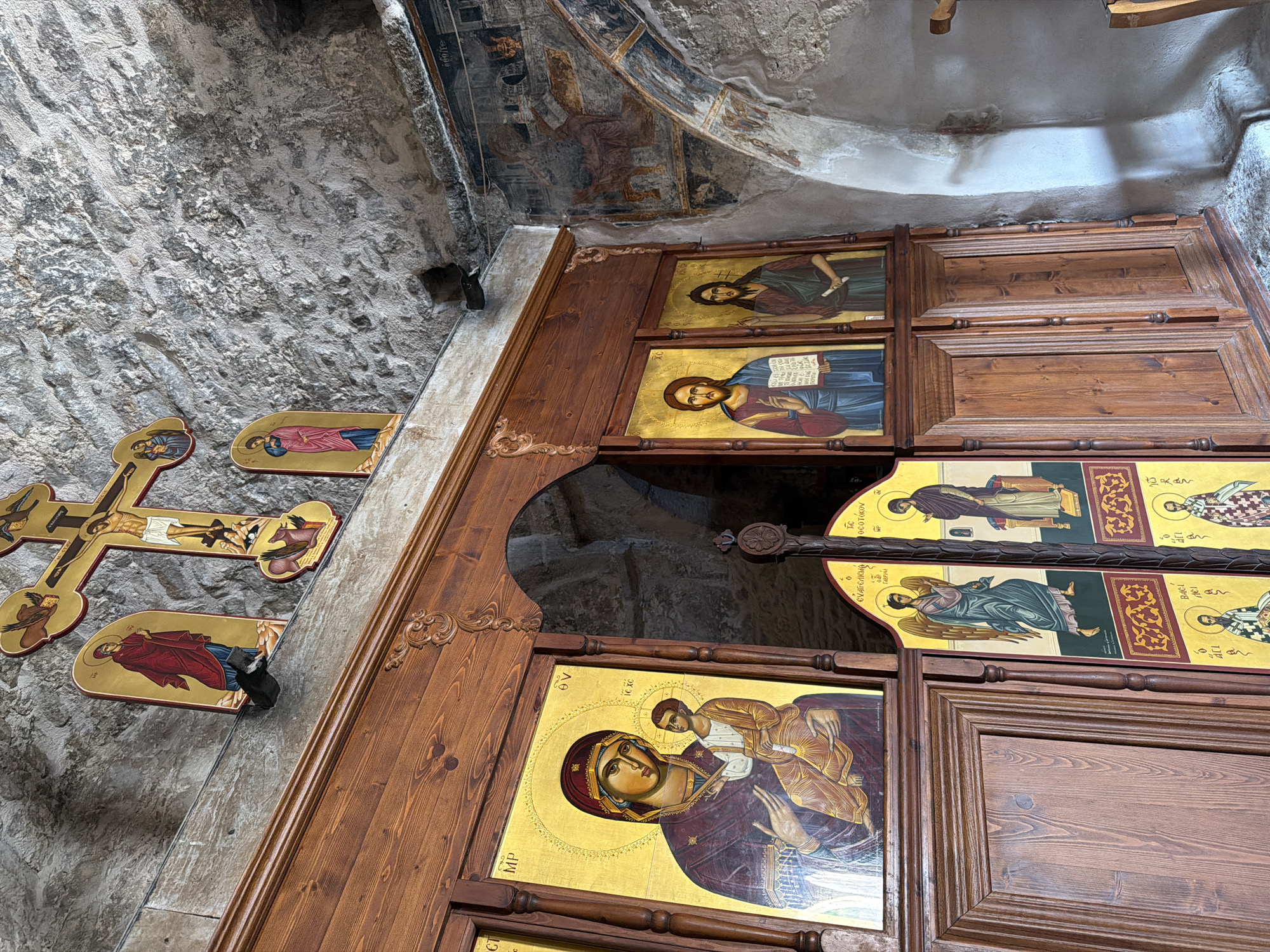The image shows an interior view of a religious building, likely a church or monastery, in Agios Therapon, Cyprus. The scene is dominated by religious iconography and architectural features.
In the foreground, a large wooden structure, possibly an iconostasis or a screen, is visible. It is adorned with several religious icons painted on gold backgrounds. The icons depict various figures, including the Virgin Mary with the infant Jesus, Jesus Christ, and other saints or religious figures, some with angelic wings. The wooden structure itself is ornately carved with decorative elements.
Above the wooden structure, a large, ornate golden cross with the crucifixion of Jesus Christ is mounted on a rough-hewn stone wall. To the left of the cross, there are smaller icons of religious figures in red and blue robes. The wall is made of grey, textured stone, suggesting an ancient or rustic construction.
To the right of the wooden structure, more religious paintings or frescoes are visible on the stone wall, arching over an opening or alcove. These frescoes appear to be faded and possibly damaged by time, depicting figures in earthy tones.
The overall environment is dimly lit, with shadows cast by the architectural elements and the objects within the space. The textures of the stone and wood are prominent, contributing to a sense of age and historical significance. The scene conveys a devotional and spiritual atmosphere.