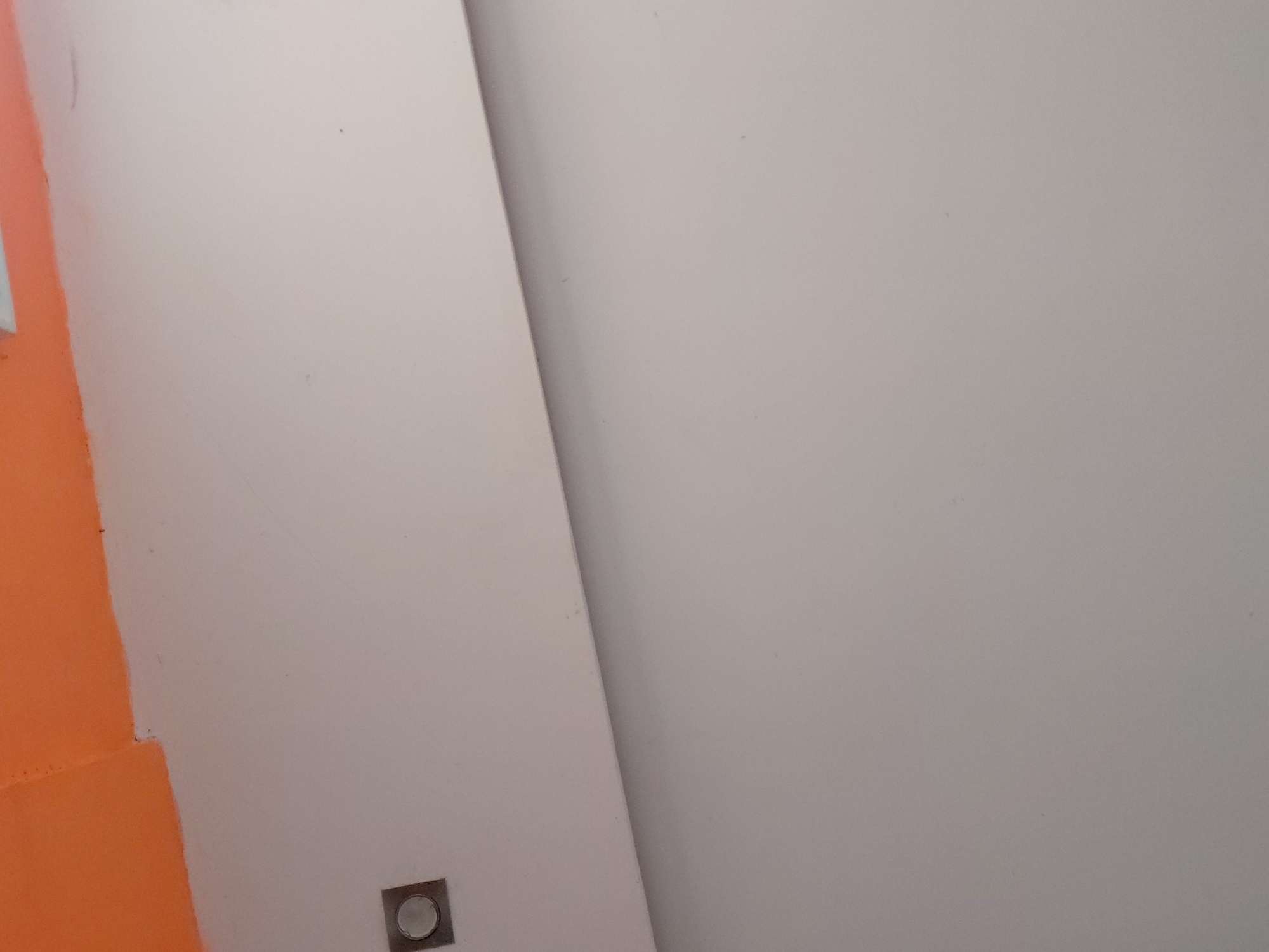 The image displays an interior architectural detail, likely a wall or a corner, characterized by a striking color contrast. A diagonal line divides the frame, separating a vibrant, bright orange painted surface on the left from a lighter, off-white or pale grey surface that dominates the right side. This line suggests a corner or a deliberate color separation on a flat wall. In the bottom left quadrant, near the intersection of the orange and lighter surfaces, a small, square-shaped fixture is visible. It appears to be a metallic or plastic cover with a circular component embedded in its center, possibly an electrical outlet, data port, or a subtle floor-level light fixture. No people or activities are present, and the scene is static. The lighting is diffused, indicating an indoor environment. There is no visible text. The location context provided is Santa Ana, Venezuela, though no specific visual cues in the image confirm this.