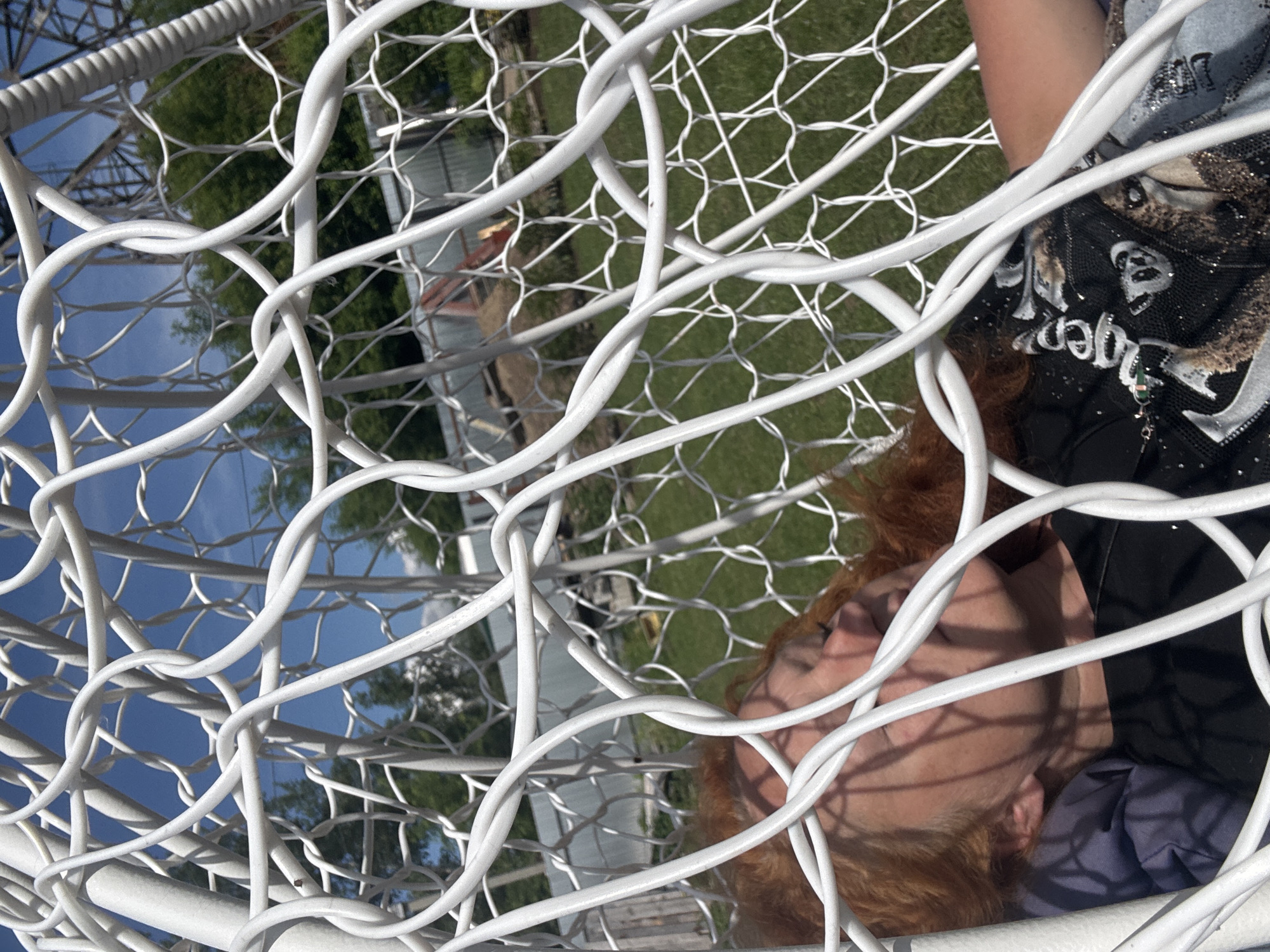The image displays a person resting outdoors, framed by a prominent white mesh structure. The subject, a person with reddish-brown hair, is lying on their side, appearing relaxed or asleep with their eyes closed. Sunlight casts distinct shadows across their face. They are wearing a dark t-shirt adorned with sparkly details, skull graphics, and white text that clearly spells out "DREAM TIGER." A dark blue fabric, possibly a pillow or part of their clothing, is visible beneath their head.
The setting is outdoors on a bright and sunny day, indicated by a clear blue sky and strong illumination. Lush green grass and foliage are visible in the background, along with parts of trees and a blurred, distant building or structure. The foreground is dominated by the white mesh, which appears to be part of a large outdoor chair or hammock. This mesh consists of thick, interwoven white strands that form an irregular, open pattern, creating a unique visual frame for the person and the natural backdrop. The overall scene suggests a moment of peaceful outdoor leisure and comfort. The specified location for this media is Chaikovsky, Russia.