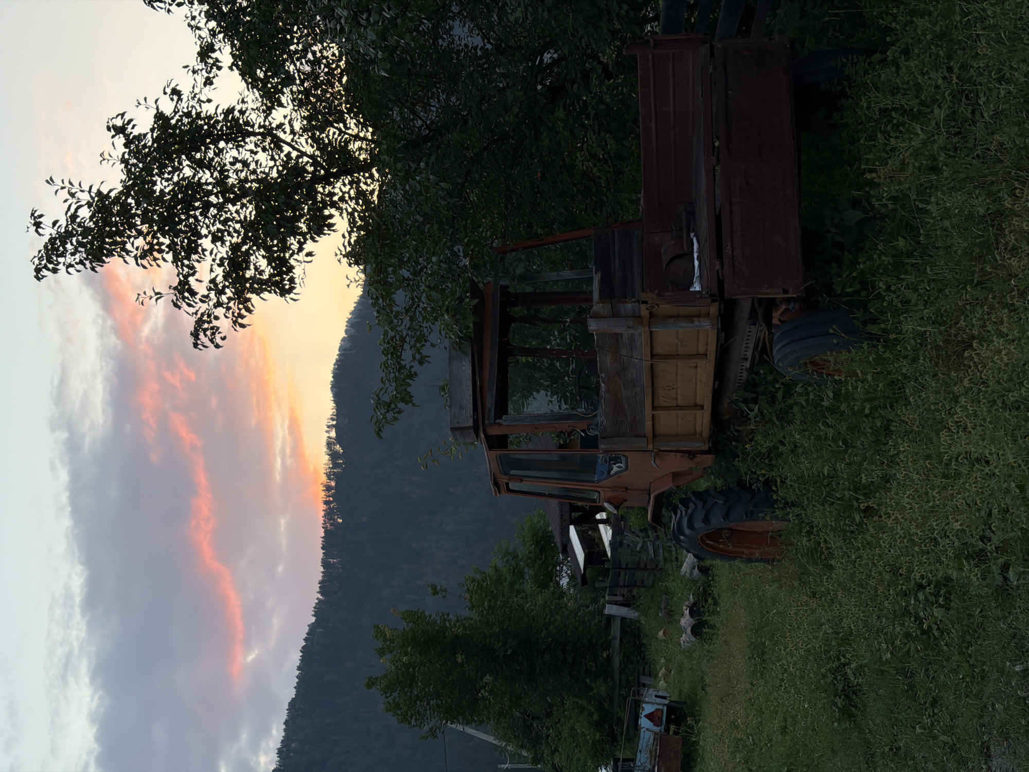In Askat, Russia, a vintage orange tractor with a wooden cargo bed sits idly in a grassy field as the sun sets behind a tree-lined mountain range. The sky is ablaze with streaks of pink and orange from the setting sun, illuminating the clouds and casting a warm glow on the scene. The tractor, appearing weathered and worn, is the central focus, surrounded by overgrown grass and vegetation. Other objects, such as a smaller trailer and some logs, are scattered around the area, suggesting a rural or agricultural setting. The overall atmosphere is one of quiet solitude and natural beauty.