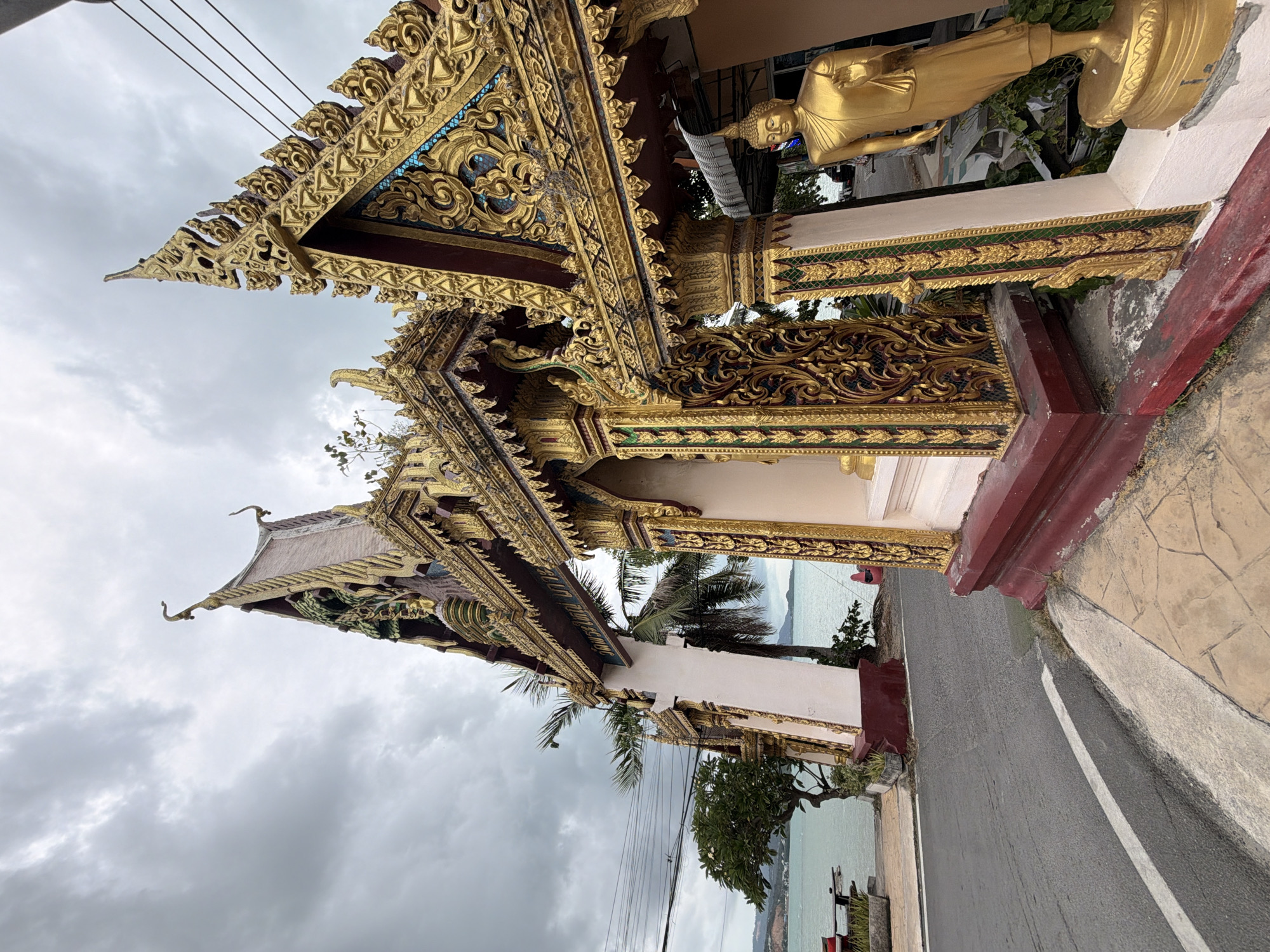 The image shows an ornate golden archway entrance to a Buddhist temple on the coast of Ko Samui District, Thailand. The archway is highly decorated with intricate carvings and gold leaf. To the right, a golden statue of a standing Buddha is visible. In the background, palm trees and the sea can be seen under a cloudy sky. A paved road runs alongside the archway.