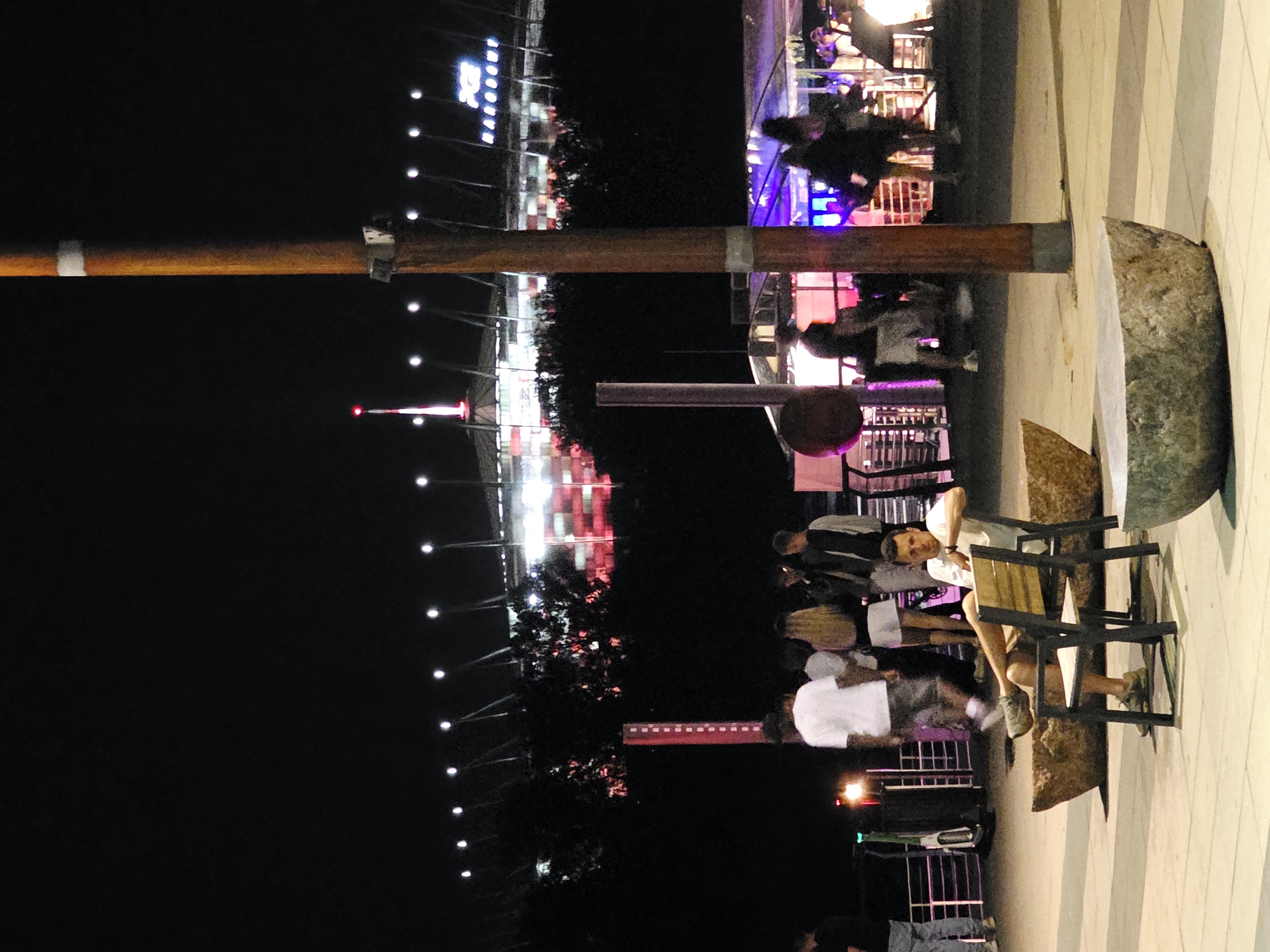 The image shows a nighttime scene in Warsaw, Poland, with a stadium in the background. A man is seated on a bench in the foreground, looking towards the viewer. He is wearing a white t-shirt and shorts, with his legs crossed and his arms resting on the chair. Behind him, a group of people are standing and walking, some under a canopy with purple lighting. The stadium in the background is lit up, with lights visible along its roofline and on the surrounding structure. The overall atmosphere is one of a public gathering or event at night.