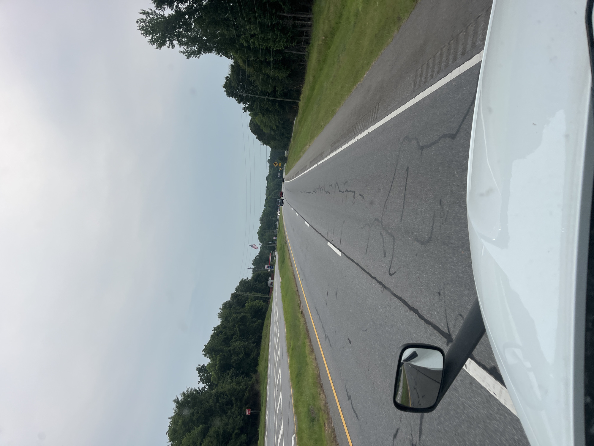 The image shows a perspective from the driver's seat of a white truck, looking forward down a two-lane highway. The road is paved and has white lane markings. There are trees lining both sides of the road. On the left side, there is a rest stop area with a sign that says "DO NOT ENTER". In the distance, there are a few cars on the road and an American flag flying. The sky is overcast. The location is Arcade, United States of America.