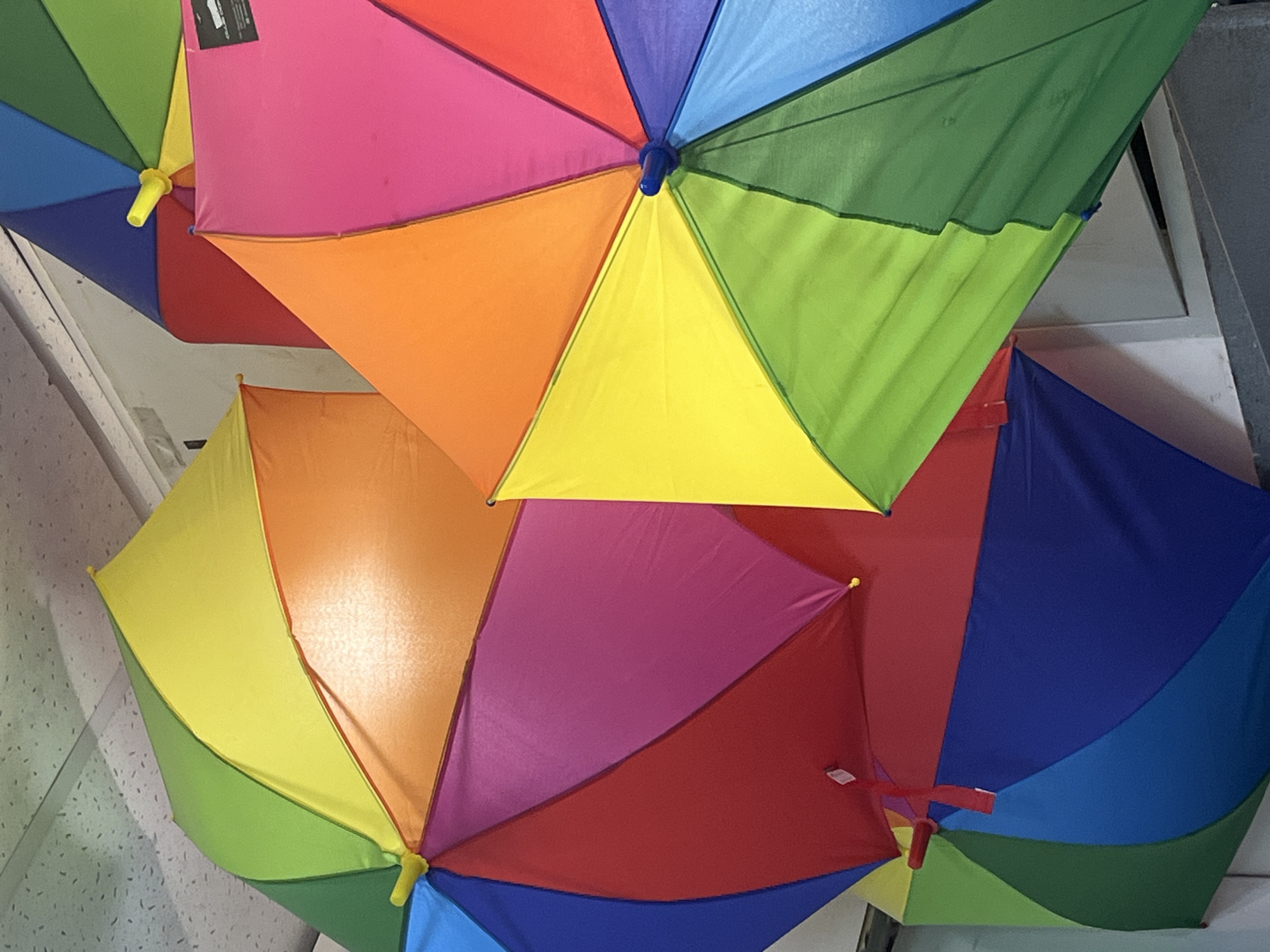 The image shows a collection of rainbow-colored umbrellas viewed from underneath. Each umbrella features segments of different colors: blue, green, yellow, orange, pink, and red. They appear to be hanging from the ceiling of a space. The scene's location in Izhevsk, Russia, is not directly discernible from the image.