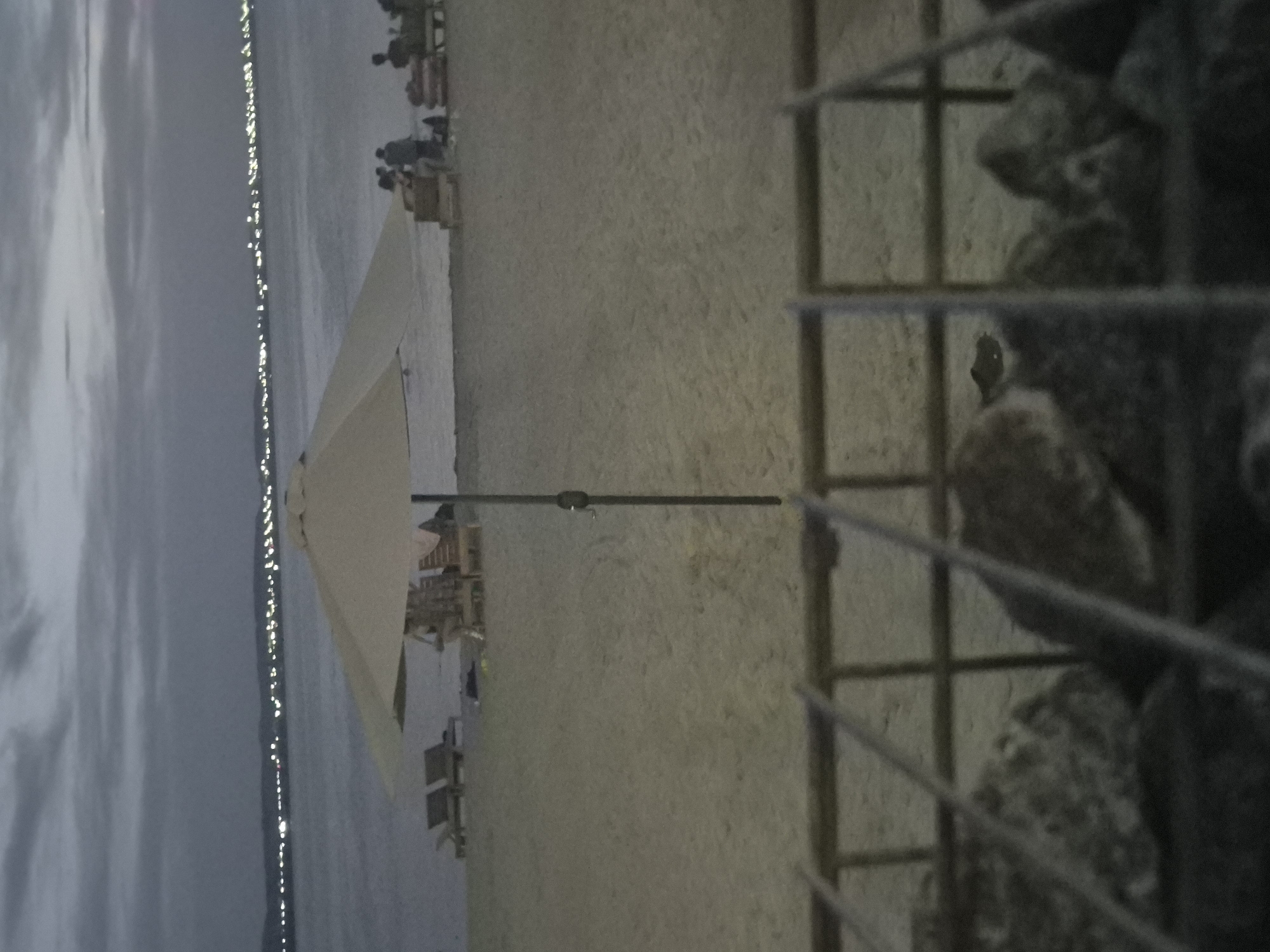 The image depicts a beach scene at dusk or dawn. The sky is overcast with muted tones of gray and blue. In the background, a distant shoreline is visible, dotted with lights that suggest a city or town. The water of a lake or sea stretches across the middle ground. On the sandy beach, several lounge chairs are arranged, some with umbrellas. A few people are visible in the distance, some appearing to be sitting or standing near the lounge chairs. A prominent beach umbrella with a light-colored canopy stands in the foreground, its pole casting a shadow on the sand. The very front of the image is obscured by a blurry foreground element, appearing to be a metal grate or fence with rocks behind it. The scene is tranquil, with a sense of quietude as day transitions to night or vice versa. This image is not from Kokshetau, Kazakhstan, as Kokshetau is a landlocked city.