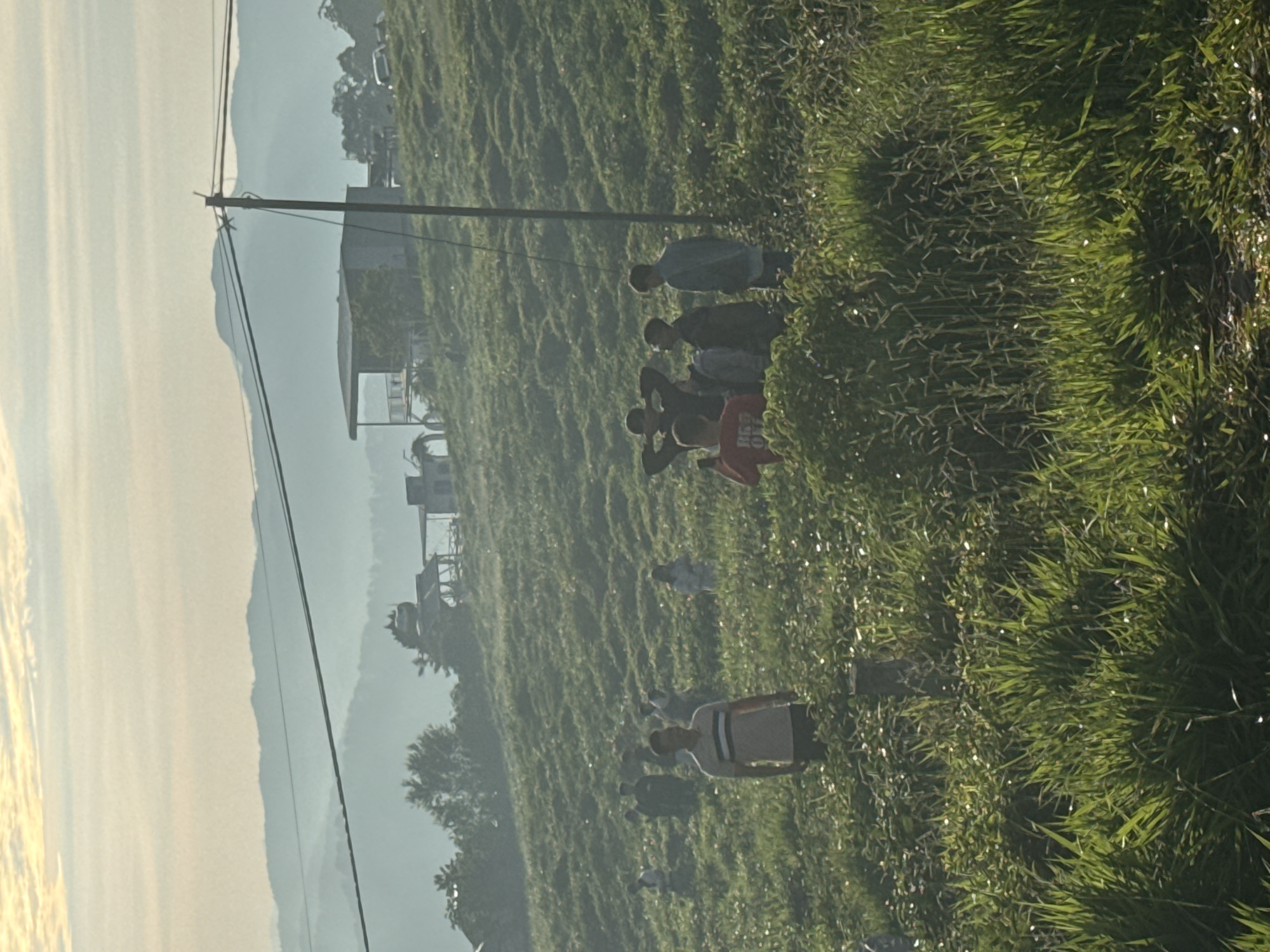 A group of people are gathered in a grassy, hilly area with scattered trees and small structures in the background, possibly in Phát Chi, Vietnam. The sun is low in the sky, casting a warm glow over the landscape and creating a hazy atmosphere with layers of distant mountains. The people, appearing to be young adults and teenagers, are spread out across the field. One man in the foreground is facing the camera, wearing a striped shirt. Other individuals are engaged in various activities, including talking, standing, and looking around. The terrain is uneven and covered in lush green grass and other vegetation. In the background, a power line runs horizontally across the frame, with a pole on the right side. Several small buildings, including one with a covered porch, are visible on the higher ground in the distance. The overall impression is a casual gathering in a natural, rural setting.