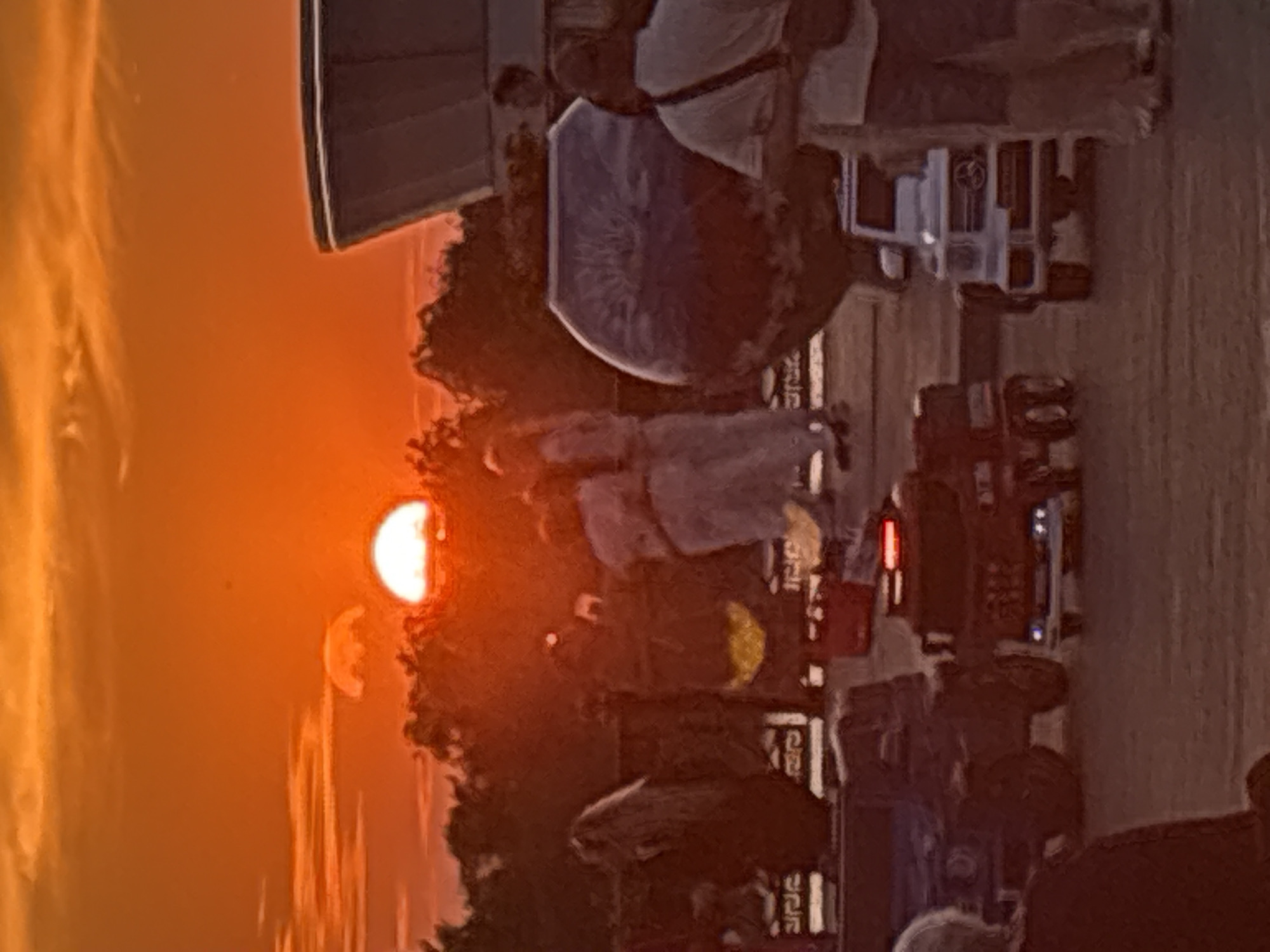 The image captures a sunset with a vibrant orange sky over a cityscape, possibly Astrakhan, Russia. In the foreground, several toy cars are visible, including a red fire truck and a white miniature Mercedes-Benz G-Wagon, suggesting a park or an area designed for children's play. Behind the toy cars, several people are standing, some appear to be adults, and a few are blurred or partially obscured. A large, decorative spherical object, possibly a sculpture or a fountain feature, is situated in the mid-ground. A building with a distinctive curved roofline is visible in the background. The overall scene suggests a recreational area or public space where families might gather during the evening.
