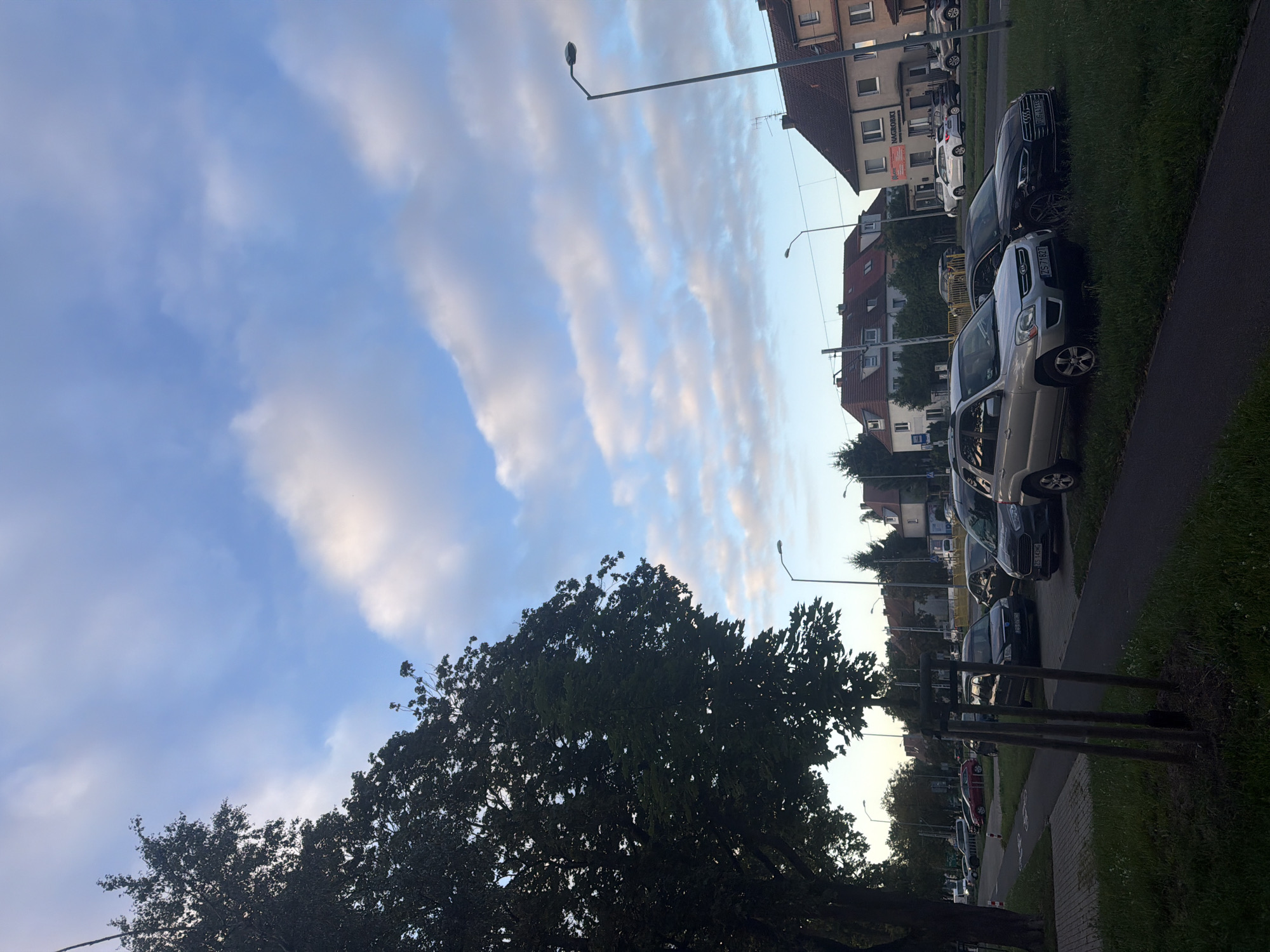 The image depicts a street scene in the city of Szczecin, Poland, likely during daylight hours with some overcast conditions. On the left, a large, leafy tree casts a shadow over a sidewalk and a paved area. Beyond this, a street is visible with several parked cars. Further in the background, residential buildings with red-tiled roofs and several stories are present, along with streetlights. One building prominently displays signage that includes "NAGROBKI". The sky above is a mix of blue and white clouds. In the foreground, a paved pathway borders a grassy area where several cars are parked. The cars include a silver SUV and a black sedan.