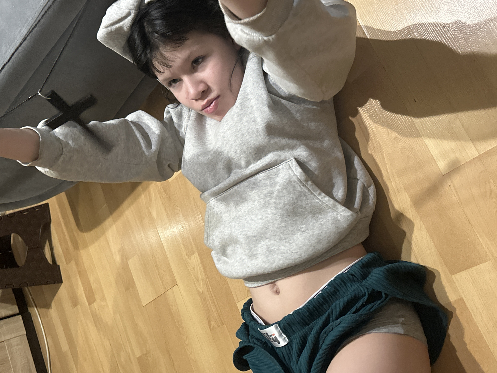 A young person is lying on a wooden floor. The individual is wearing a gray hooded sweatshirt and green shorts. The subject is holding a cross necklace in their right hand, which is extended to the left of the image. Part of a gray object is visible in the upper-left of the image, and a brown object is visible in the lower-left of the image. The subject's abdomen is visible. The image is taken from above the subject. The location is not discernible from the image.