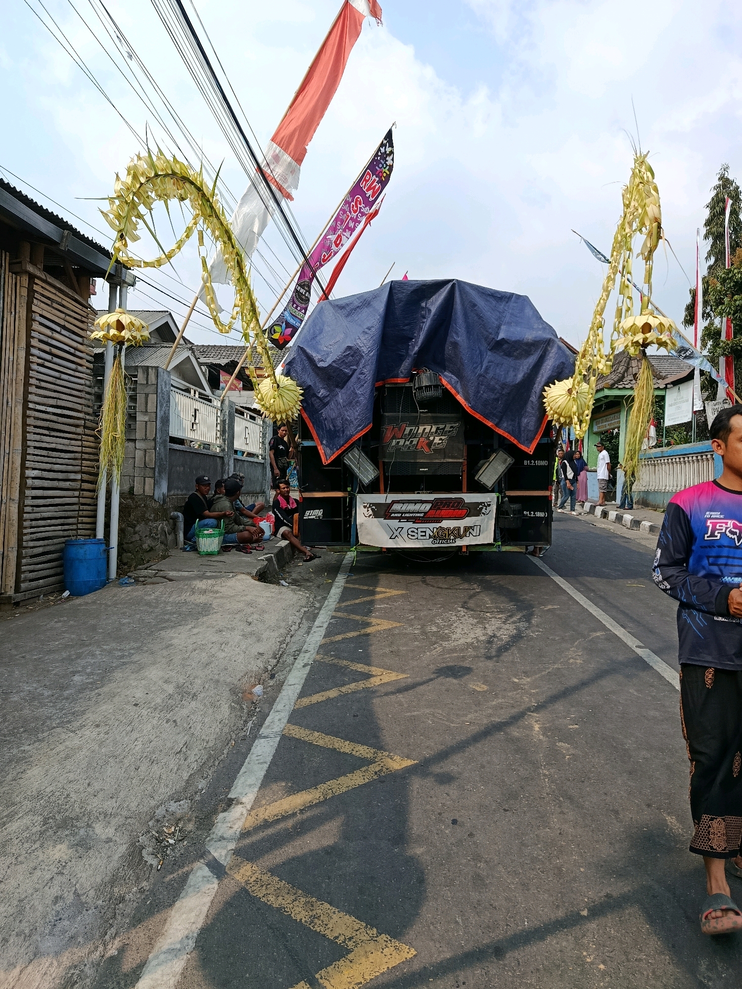 The image depicts a street scene in Gubukklakah, Indonesia, under a bright sky with scattered clouds. In the foreground, a man wearing a colorful long-sleeved shirt and patterned pants stands facing to the right. Behind him, a truck is parked on the side of the road, covered by a large blue tarp. The truck appears to be a sound system setup, with speakers visible on the back. Decorative elements, possibly made from palm leaves, are arranged on either side of the truck, arching upwards and adorned with yellow, conical structures. Several people are seated on the ground and on a low wall to the left of the truck, seemingly engaged in conversation or resting. A blue plastic barrel is situated near them. The road has painted yellow markings. In the background, there are houses, fences, and trees, suggesting a residential area. Indonesian flags and other banners are also visible, indicating a possible festive occasion or event.