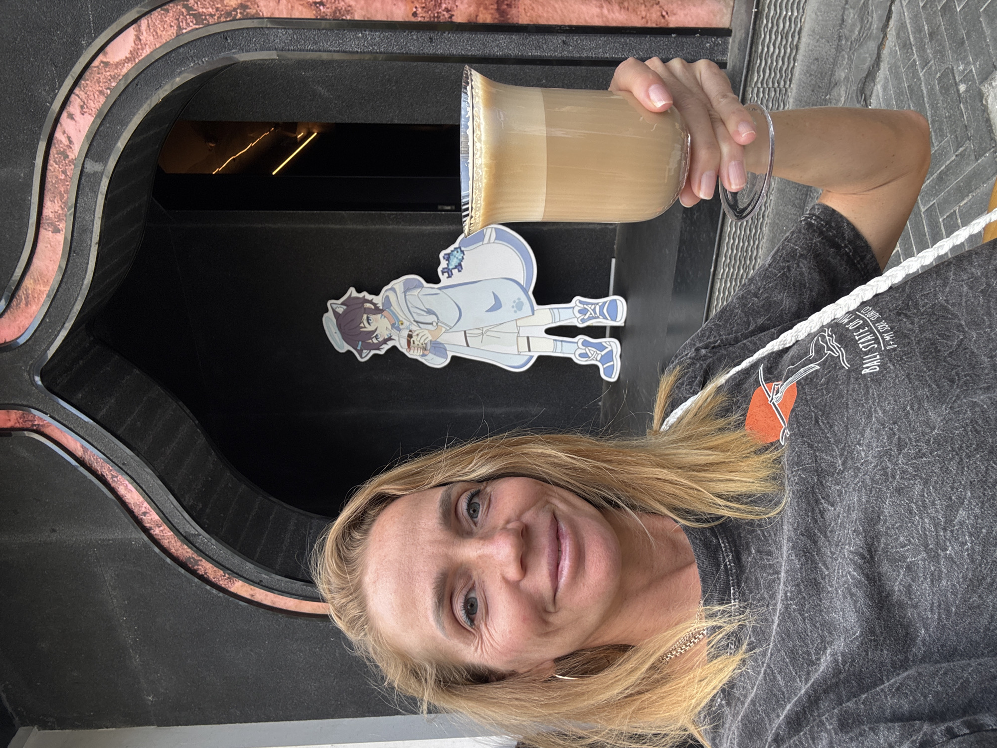 A woman with fair skin, blond hair, and wearing a gray t-shirt is holding a glass of coffee in front of a black doorway with an arched frame. Attached to the door, behind the glass, is a cartoon anime figure. The location is Jinjiang District, China.