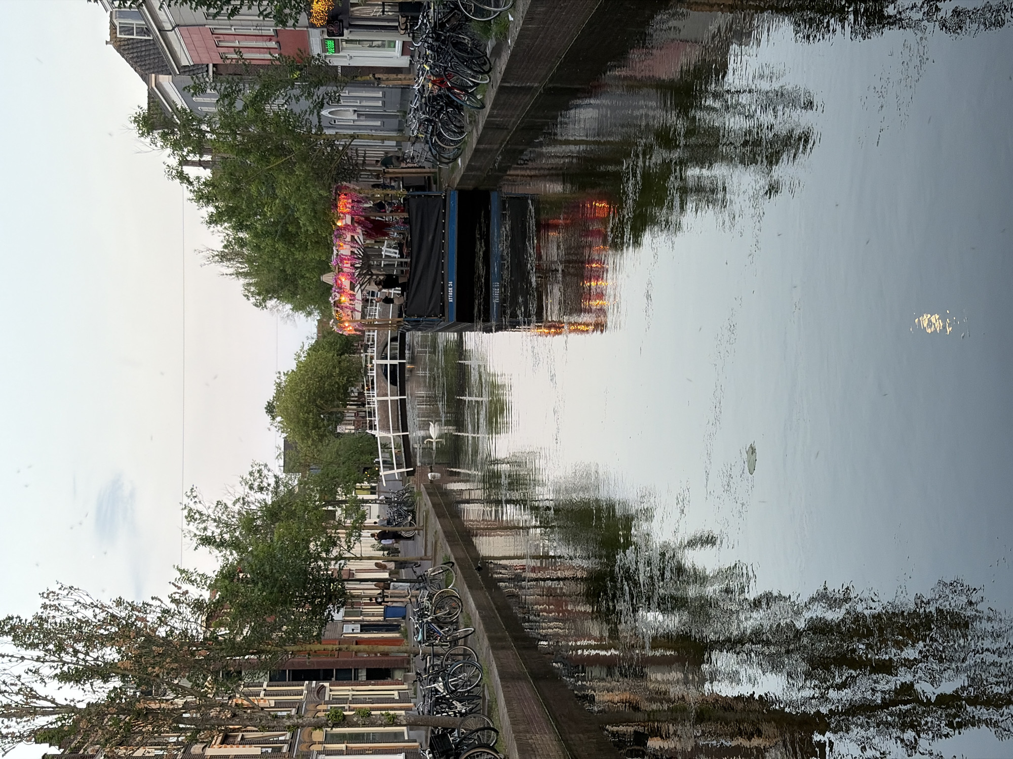 The image shows a canal in Delft, Netherlands. A barge-like restaurant or bar floats on the canal, decorated with pink floral arrangements and lights. People are seated at tables on the barge. The buildings along the canal bank are typical Dutch row houses, with bicycles parked along the side of the canal. The trees are reflected in the water. The sky is overcast. The scene appears to be taken during the evening.