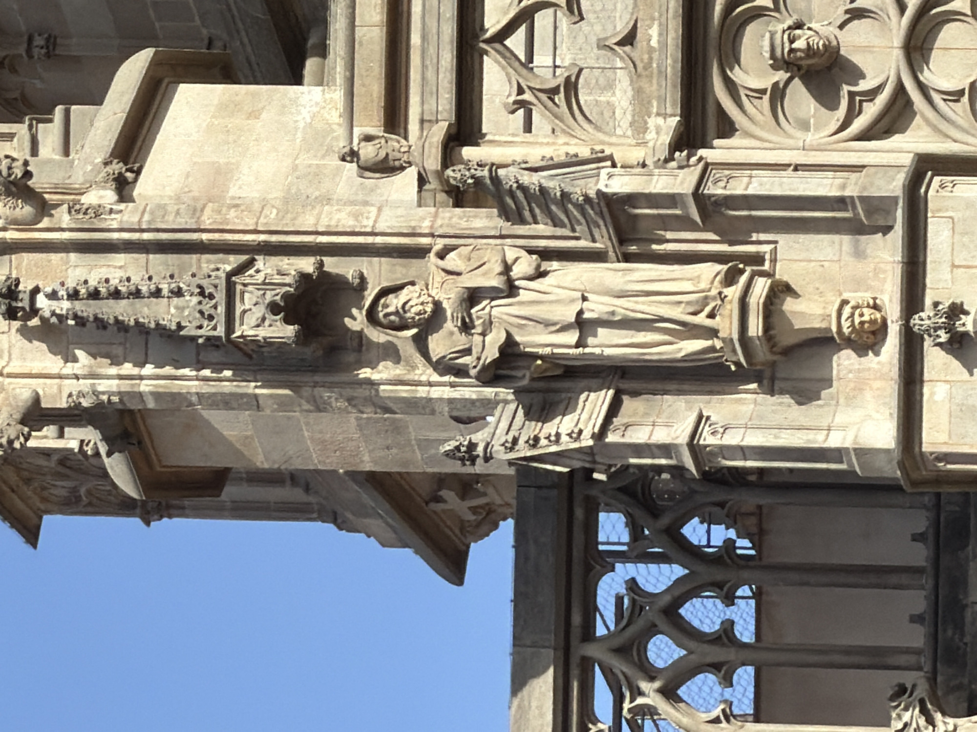 This is a close-up, low-angle, vertical shot of ornate stone architectural details on the exterior of a building, likely a cathedral, under a clear, bright blue sky. The stone is a light beige or tan color and shows signs of age and weathering.
The most prominent subject is a statue of a robed figure lying down, with a hood covering their head. The figure appears to be a saint or monk, with carved folds in the robe and hands resting on the chest. Below this statue, another carved figure with a beard and similar robes is visible. Above the reclining statue, there are decorative architectural elements, including carved faces and intricate geometric patterns within circular frames.
Below the main statues, there's a prominent carved ledge or balcony featuring a detailed frieze with repeating motifs and a central decorative element. Further down, a section of a Gothic-style window with intricate tracery is visible, framing the bright sky. Small, grotesque carved figures, possibly gargoyles or decorative corbels, are present at various points along the architecture.
The lighting suggests it is daytime, likely morning or afternoon, with strong sunlight casting shadows that emphasize the depth and detail of the carvings. The clear blue sky indicates fair weather. The style of the architecture, particularly the Gothic tracery and detailed stone carving, points to a European location, consistent with Barcelona, Spain. There is no visible text in the image.