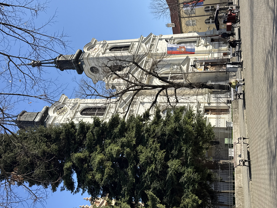 The image depicts the Cathedral Church of Saint Nicholas, located in the city of Sremski Karlovci Municipality, Serbia, on a clear, sunny day. The dominant structure is a large, white, baroque-style church with two symmetrical bell towers topped with dark, ornate spires and featuring visible clock faces. The church facade is detailed, with arched windows and decorative elements. A vertical Serbian national flag (red, blue, white tricolor with coat of arms) is prominently displayed on the right side of the church's entrance area.
In the foreground, a public plaza with a patterned paver surface extends. A large, dark green conifer stands on the left, partially obscuring the left church tower. A leafless deciduous tree is positioned in front of the church's central facade. Multiple dark-colored park benches with wooden slats are arranged on the plaza.
Three individuals are visible on the plaza. One person, wearing a light-colored top and dark trousers, is seated on a bench on the right side of the frame. Another person in dark clothing is seated on a bench further right. A third person, also in dark clothing, is standing near a red, vintage-style three-wheeled vehicle with a canopy, parked on the far right. A fourth individual is partially visible walking near the vehicle. A dark metal fence runs along the base of the church building, separating it from the plaza. The scene is illuminated by strong natural light, casting distinct shadows.