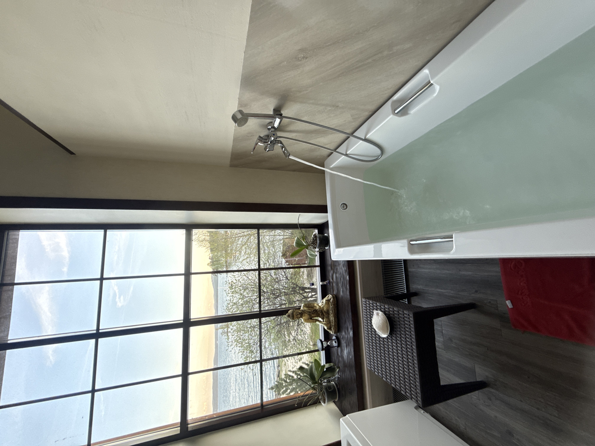The image shows a bathroom in Zolotoi Plyazh, Russia, featuring a large window offering a view of a beach and the sea. The window has a black frame divided into multiple panes. A white bathtub is positioned near the window, with water flowing from a chrome faucet. On the window sill, there is a golden Buddha statue and two potted plants. A small black wicker table with a white object on it stands next to the tub, and a red towel is visible nearby on the dark-grey wooden floor. The room's ceiling is painted in two different tones: a light beige and a greyish-beige.