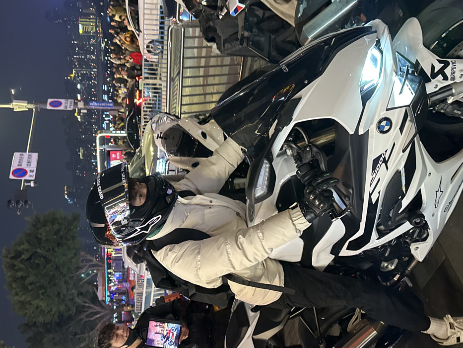 This nighttime image captures an urban scene in Yuzhong District, China. In the foreground, a person is seated on a white and black BMW S1000RR sport motorcycle, facing slightly right. The individual wears a black full-face motorcycle helmet with a reflective visor displaying "GIVES YOU WINGS" and "LOTUS P" text, a black balaclava, a cream-colored puffer jacket, black pants, and white sneakers. Black motorcycle gloves are worn, with hands gripping the handlebars. A black backpack is on the individual's back, and a light-colored crossbody bag is slung over the shoulder. The motorcycle displays BMW, "S1000RR," and Alpinestars logos, with an "Akrapovic" sticker visible on the lower front fairing. The motorcycle's headlights are illuminated.
To the immediate left, a second male individual with dark hair, wearing a black jacket and a black face mask pulled down below the chin, smiles slightly while holding a tablet displaying an image of a motorcycle. A metal barrier separates the foreground motorcycles from a bustling mid-ground street and pedestrian area. Numerous indistinct people populate the mid-ground promenade, and several cars are visible on the street beyond the barrier. In the distant background, a brightly lit city skyline with multiple buildings extends across what appears to be a body of water, indicative of a riverine city, consistent with Yuzhong District. Overhead, a utility pole with traffic lights and Chinese signage, including a "禁止鸣笛" (No Honking) symbol and time restrictions, is visible. The scene is illuminated by artificial street and building lights.