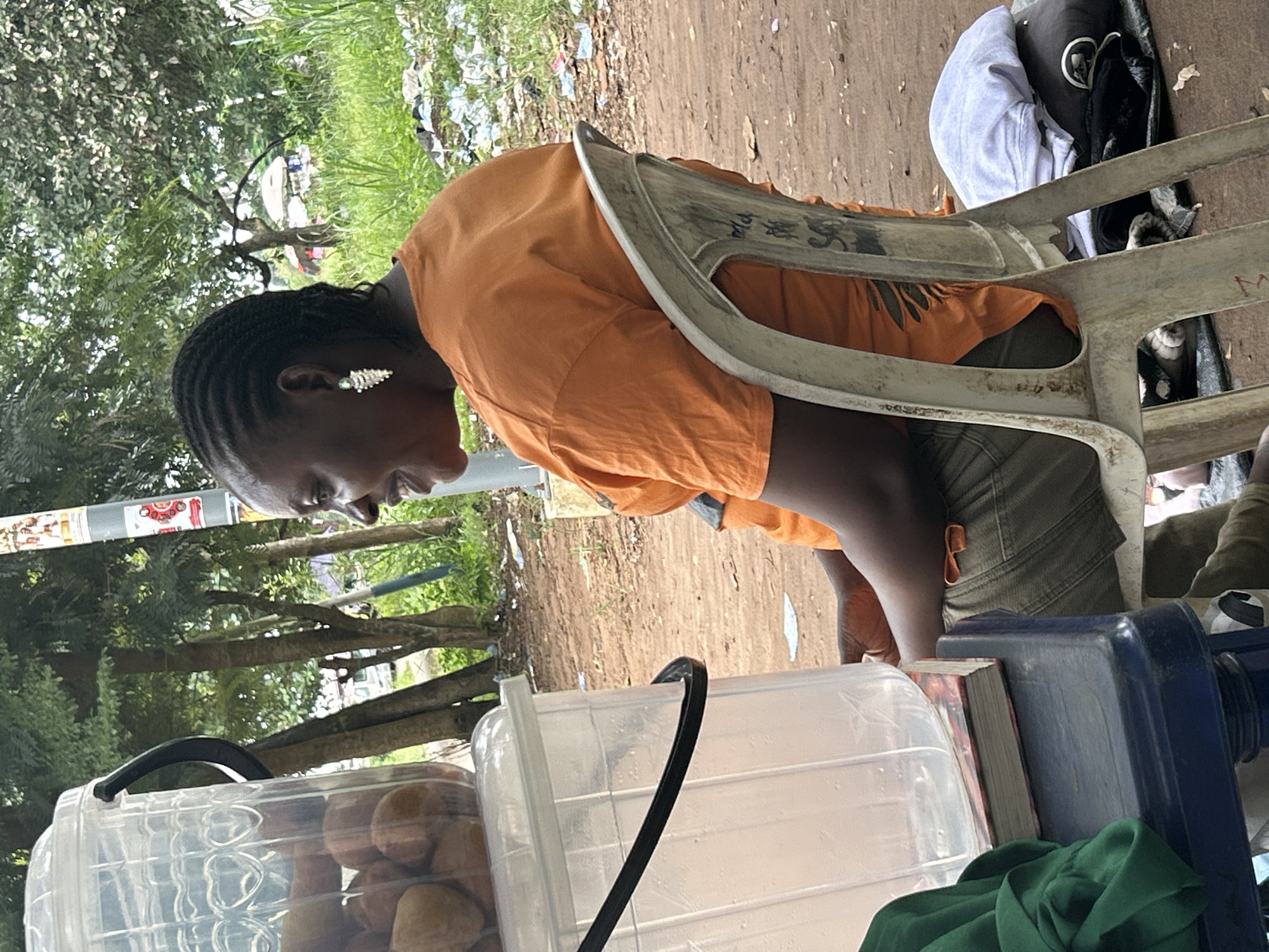 In Ihiagwa, Nigeria, a woman is seated in a white plastic chair, her profile visible as she looks to her right. She is wearing an orange t-shirt and green pants, adorned with large, sparkling earrings. Beside her, a clear plastic container filled with round, brown objects is perched on a blue surface. The background reveals a dirt path, sparse vegetation, and trees. The scene appears to be outdoors, possibly at a market or roadside stall, given the presence of bundled items and scattered debris in the background.