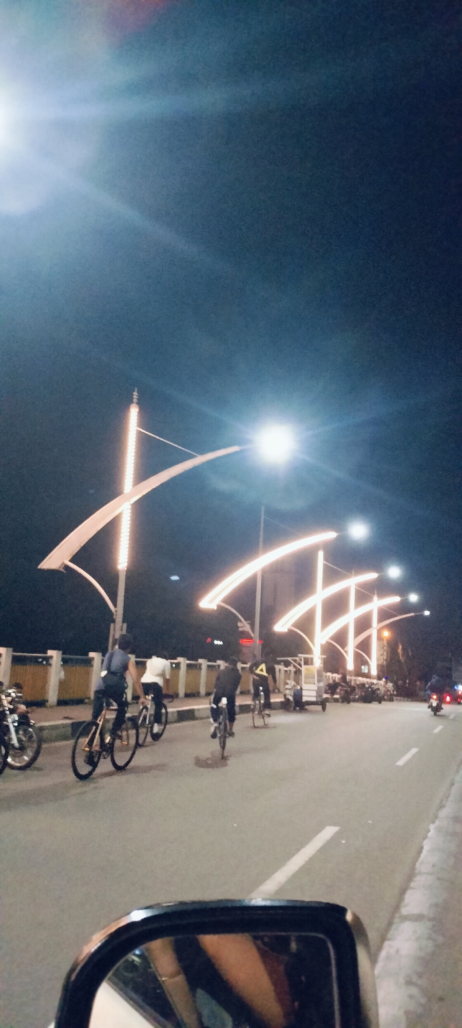 The image depicts a vibrant night scene on a bridge or elevated road in Banda Aceh, Indonesia. Several people are actively cycling along the asphalt road, some individually and others in small groups, suggesting a recreational evening ride or commute. The perspective is from inside a vehicle, with a portion of a rearview mirror visible in the bottom left corner.
The scene is brightly lit by decorative streetlights, which are a prominent feature. These unique lights consist of tall poles with multiple arched, illuminated sections that crisscross above the road, casting a warm, yellowish-orange glow. A low railing or barrier runs along the left side of the road, indicating the edge of the bridge structure.
Besides the cyclists, other vehicles are present: several motorcycles are parked along the left, and a three-wheeled vehicle (possibly a "becak" or similar local transport) is parked on the right side of the road. Another motorcycle can be seen further down the road on the right. The road itself has clear white lane markings.
The time of day is clearly night, with a dark sky contrasting sharply with the bright artificial illumination from the streetlights, which also produce visible lens flares. The weather appears clear. The atmosphere is lively and well-lit, with people enjoying the cool evening air. There is no discernible text visible within the image.