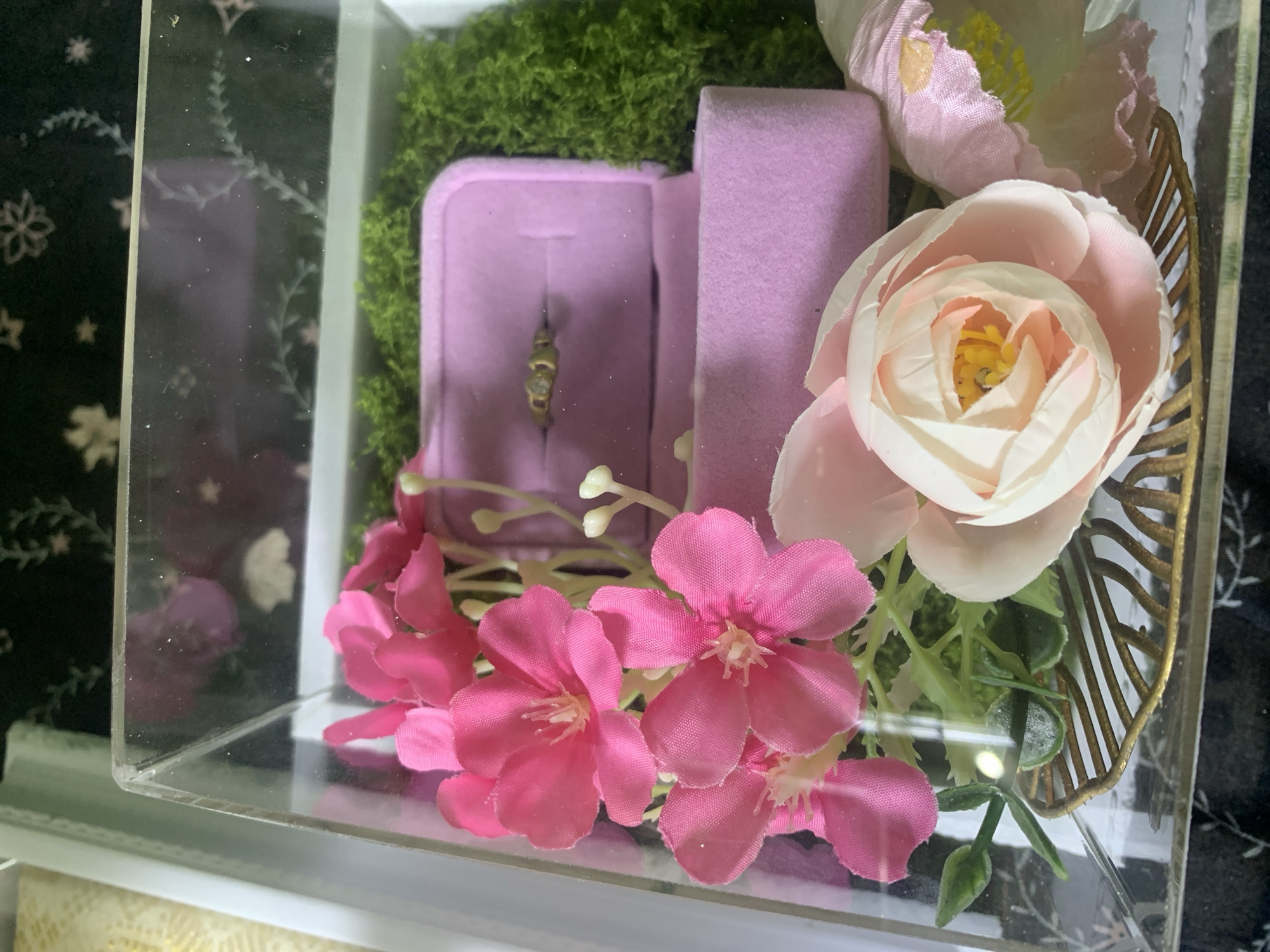 This media file is a close-up shot of a clear acrylic box containing decorative items, likely for a proposal or wedding. The main subject is a ring box, open to reveal a delicate gold ring with small stones. Surrounding the ring box is an assortment of artificial flowers in shades of pink and white, along with green moss. A gold-colored decorative leaf element is visible on the right side of the arrangement. The overall aesthetic is romantic and celebratory. The setting appears to be indoors, given the lighting and the lack of natural elements beyond the decorative moss and flowers. There is no visible text in the image. The focus is on the ring and the floral arrangement, suggesting a moment of anticipation or presentation of a significant gift. The location context provided, Kuala Pembuang, Indonesia, is not visually evident in the image itself.