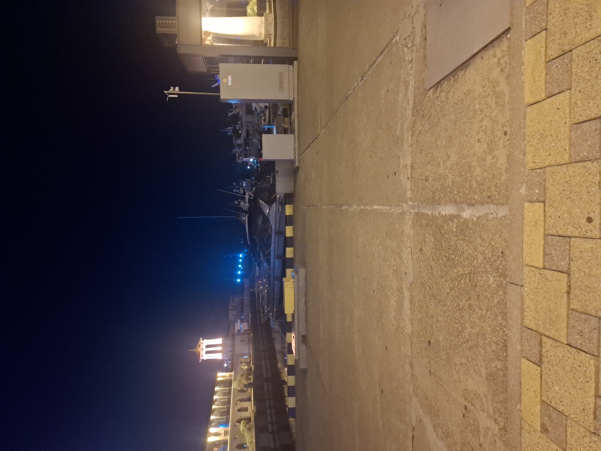 A nighttime view of a marina in Sochi, Russia. In the foreground, a concrete walkway is visible, bordered by decorative yellow and brown paving stones. Beyond the walkway is the water of the marina, with several boats moored. A striped yellow and black barrier runs along the edge of the marina. In the background, a building with arched windows and a tower lit with white lights is visible on the left. On the right, there are industrial-looking structures, including grey electrical cabinets and a building with large windows, illuminated from within. The sky is dark blue, suggesting it is night.