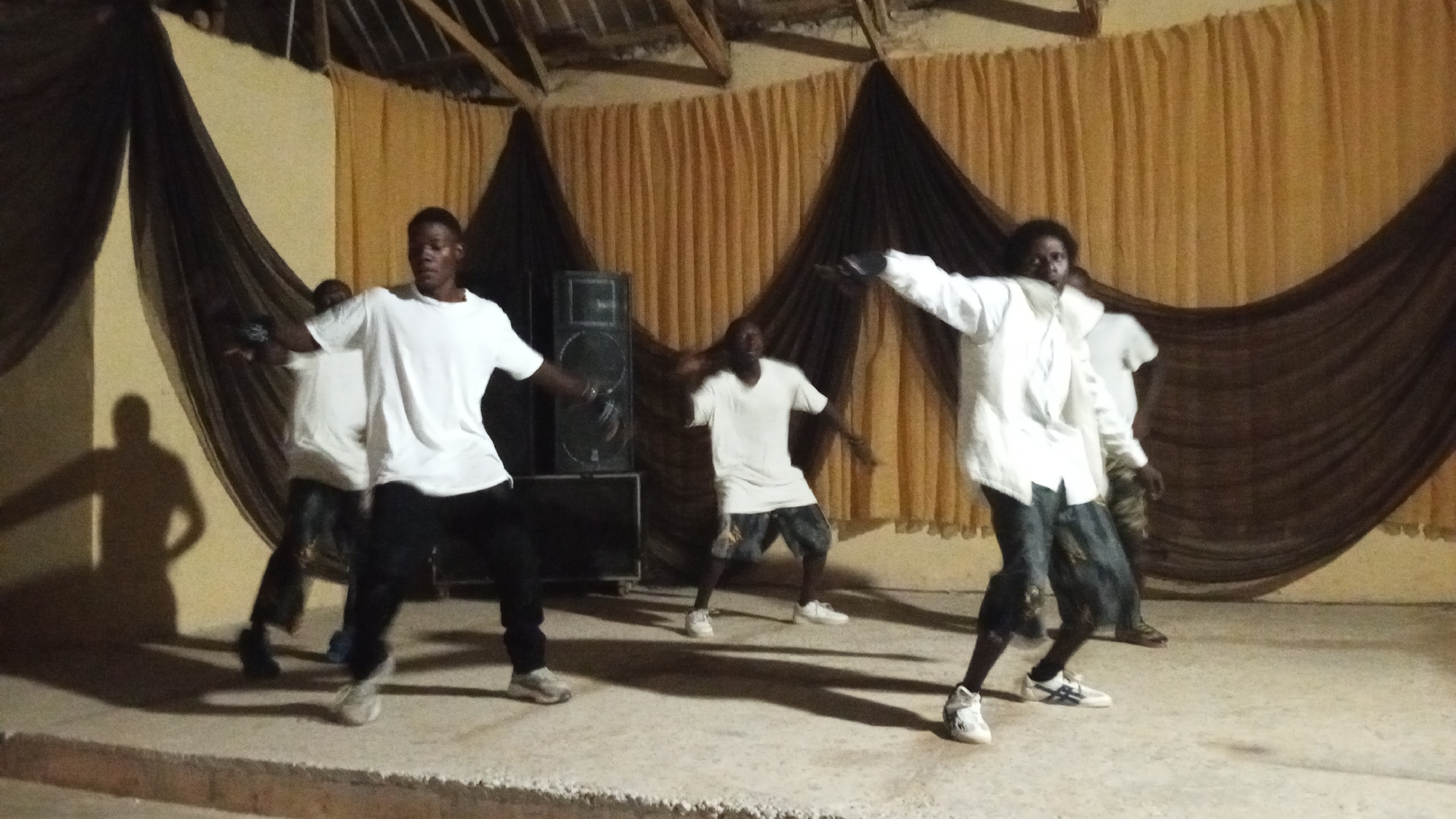 A group of five young men are performing on a stage. They are all wearing light-colored shirts and dark pants, with the man on the right also wearing a white button-down shirt. The stage is decorated with light brown and dark brown drapery. A speaker is visible behind the dancers. The location appears to be indoors under a roof. The city and specific location are not discernible.