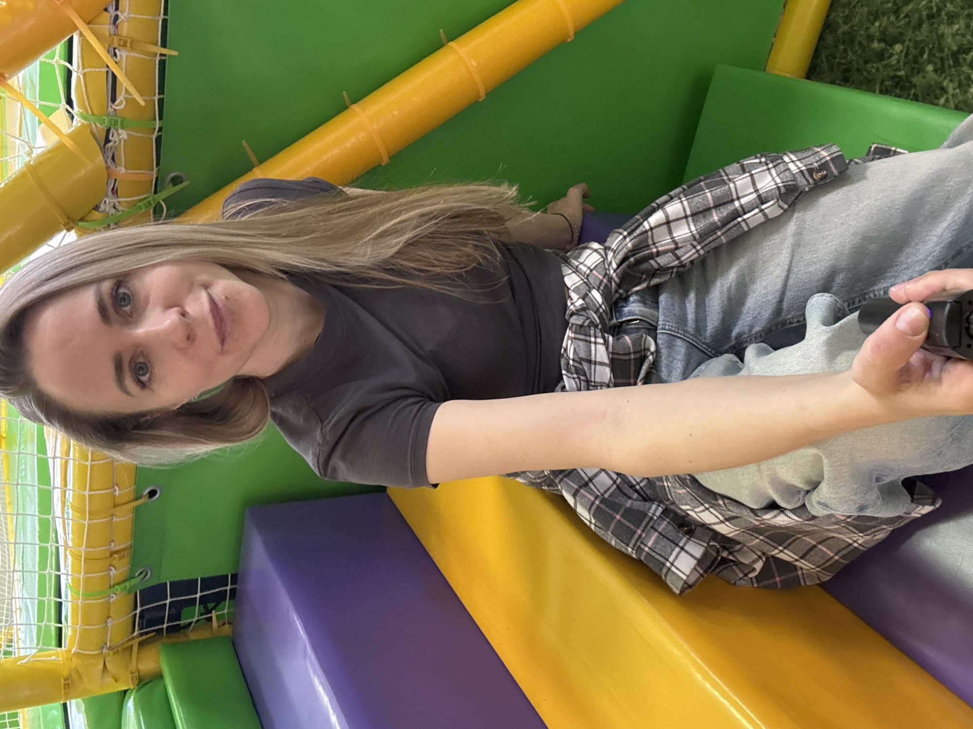 A woman is posing for a selfie in what appears to be an indoor play area with colorful structures, tubes, and netting. She has blonde hair and is wearing a dark gray t-shirt, jeans, and a plaid shirt tied around her waist. She is holding an electronic device in her outstretched hand. The play area consists of green, yellow, purple, and orange soft play equipment. The specific location in the city of Chaikovsky, Russia is not discernible from this single image.