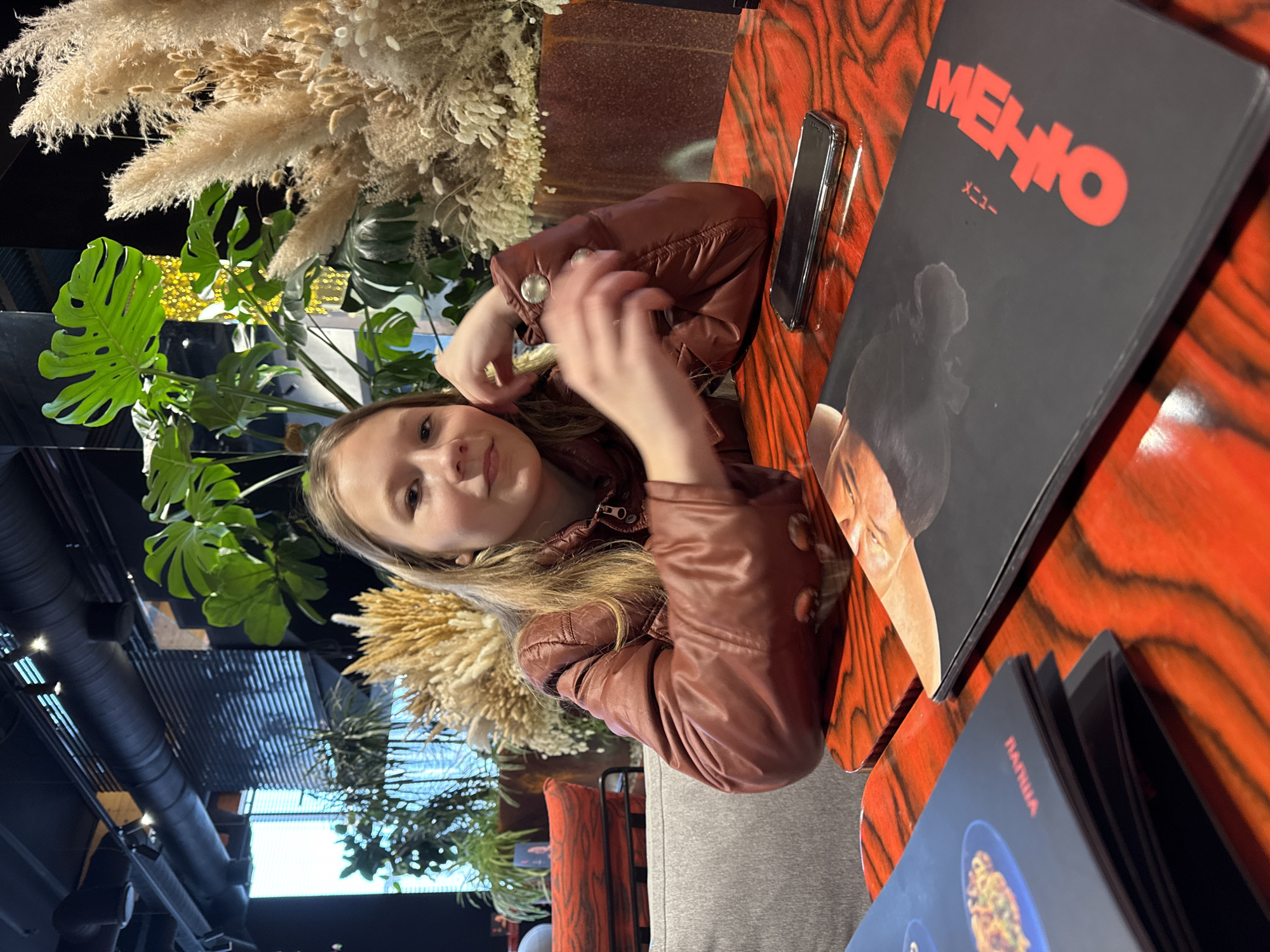 A girl with light brown hair wearing a brown jacket is seated at a restaurant table. She is looking at the camera, holding her hands near her face. A black cell phone is on the table next to her. A menu with the word "МЕНЮ" in red is also on the table, alongside another menu with the word "ПАПША" written on it. The table has a reddish-brown wood grain pattern. In the background, there are decorative plants, including pampas grass and monstera leaves. The location is Rostov-on-Don, Russia.