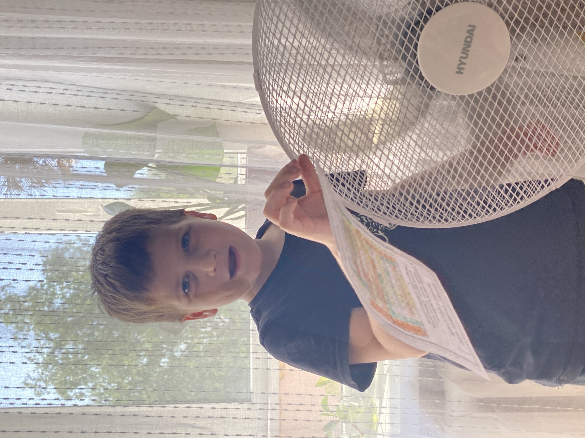 A child is holding a piece of paper near a white Hyundai fan in front of a window. Outside the window, trees are visible. The location is likely indoors, given the presence of the fan and the curtains. The city of Chaikovsky, Russia, cannot be confirmed or denied from the image alone.