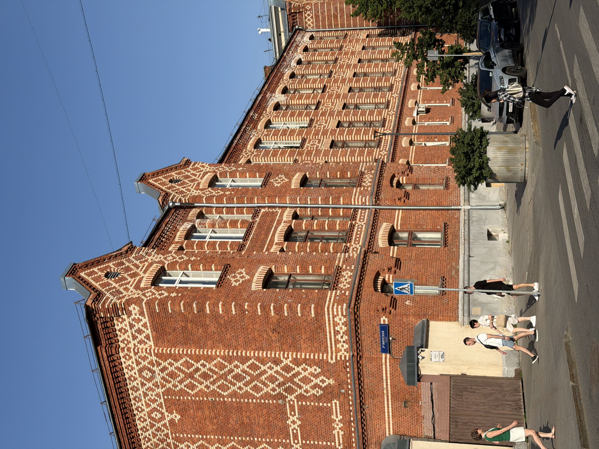 The image shows a three-story red brick building in Moscow, Russia. The building has distinctive white brickwork patterns and multiple windows on each floor. A street sign for 2nd Miusskaya Street is visible. Several people are walking on the sidewalk. Parked cars are present along the street. The sky is clear and blue.
