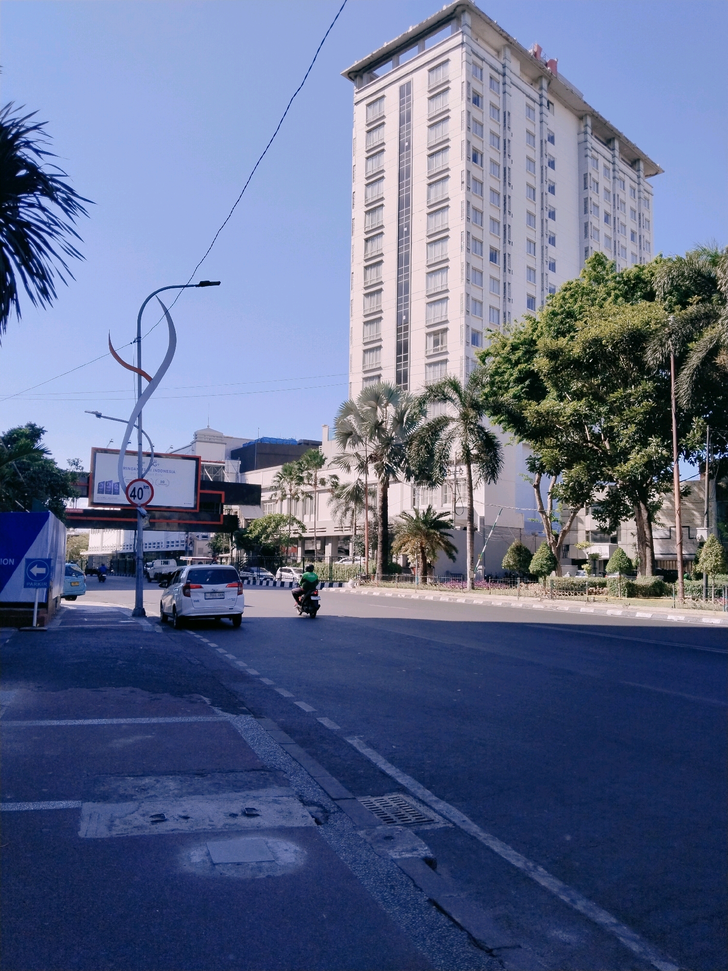 This is a medium-shot, horizontal, outdoor, daytime, eye-level photograph taken in Surabaya, Indonesia. The image features a tall, multi-story white building with many windows, possibly a hotel or office building, dominating the background. In the foreground, a two-lane asphalt road runs horizontally across the scene. A white SUV is parked in the right lane, and a person on a motorcycle is riding in the left lane, moving away from the viewer. A pedestrian sidewalk is visible on the left side of the road, with a blue sign with white lettering that appears to be a directional sign. A tall pole with a circular speed limit sign indicating "40" and an advertisement billboard are positioned near the sidewalk. Lush green trees, including palm trees, are scattered throughout the scene, some obscuring parts of the buildings. The sky is a clear, light blue. The overall scene depicts a typical urban streetscape in Surabaya, with vehicles, buildings, and vegetation under bright daylight conditions.