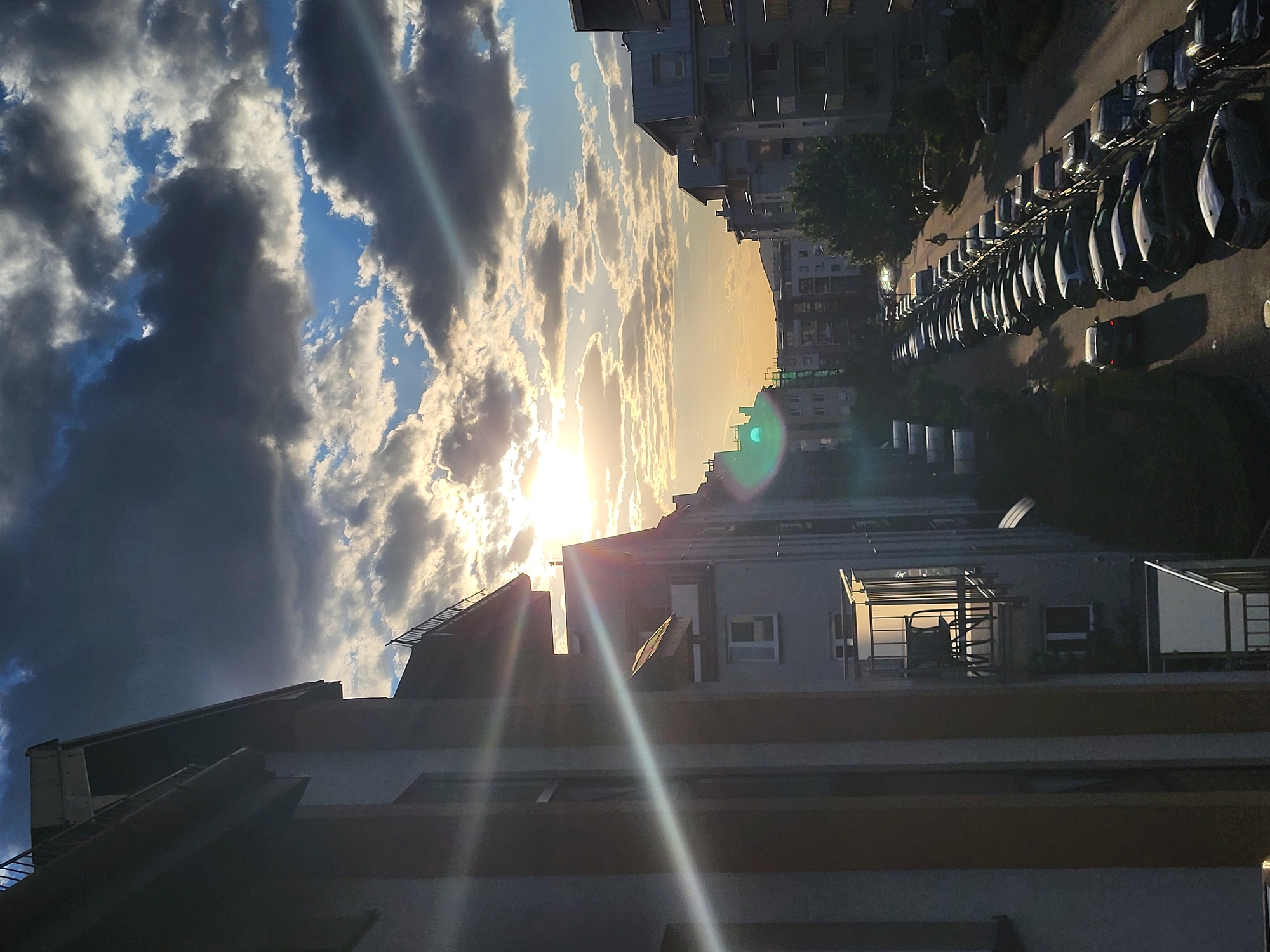A low-angle, medium shot depicts a city scene at dusk or dawn, with a blend of natural and artificial light sources. The sky is filled with a dramatic cloudscape ranging from dark grey to light white, reflecting the sun's rays, which create flares in the camera lens. Below the sky, the tops of tall buildings are visible. The buildings appear to be made of concrete and feature rows of windows and balconies. A long line of parked cars stretches down the right side of the image, indicating a residential or commercial area. The overall atmosphere is urban and dynamic, capturing the transition between day and night. The location appears to be a densely populated area, but it is not possible to determine the precise location.