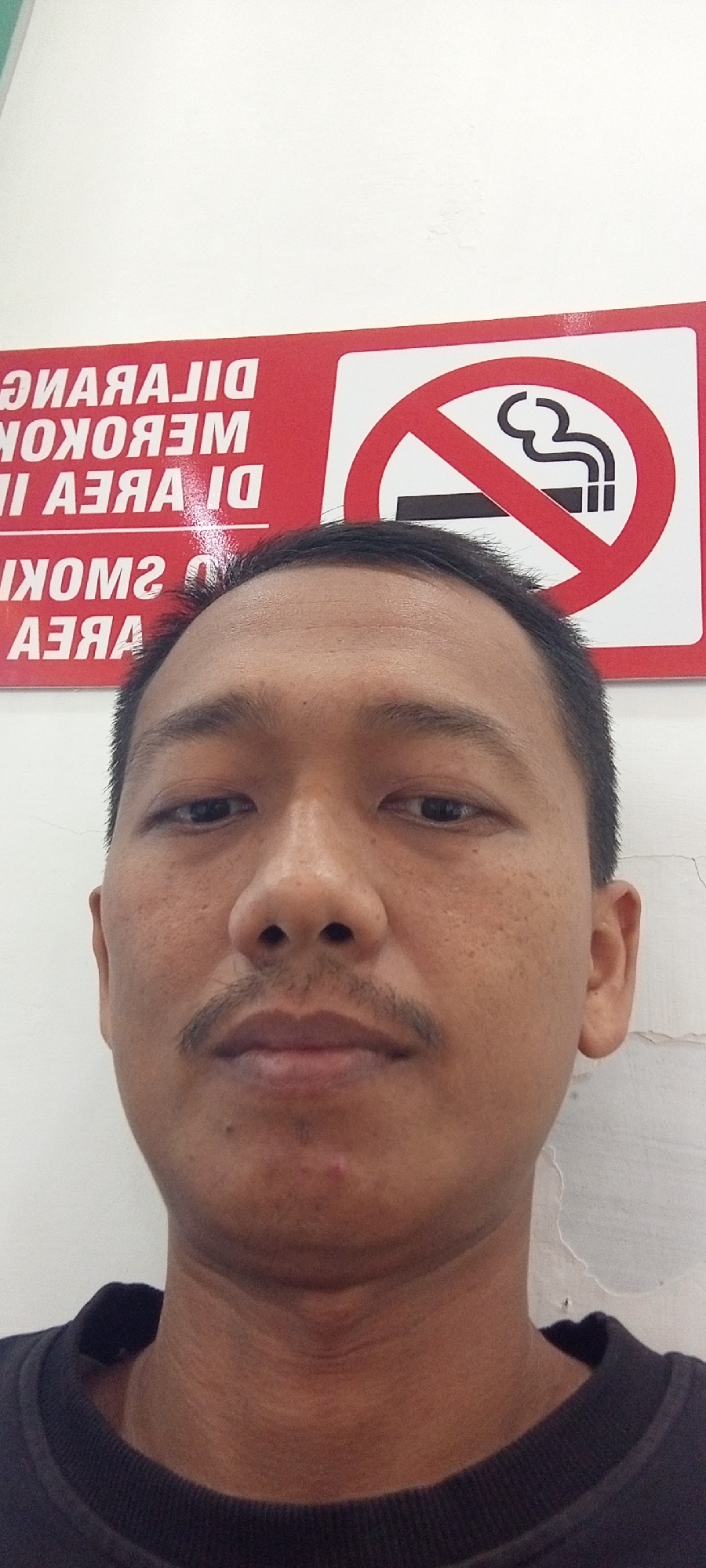 A close-up selfie features a man with short black hair and a mustache. He is wearing a black crew neck shirt. Behind him, a red and white sign with white text reads "DILARANG MEROKOK DI AREA II" and "NO SMOKING AREA". The sign also includes a red circle with a diagonal line through it, symbolizing prohibition, and an image of a lit cigarette emitting smoke. The background appears to be a white wall with some minor cracks and imperfections. The image was taken in Lhokseumawe, Indonesia, at a location where smoking is prohibited.