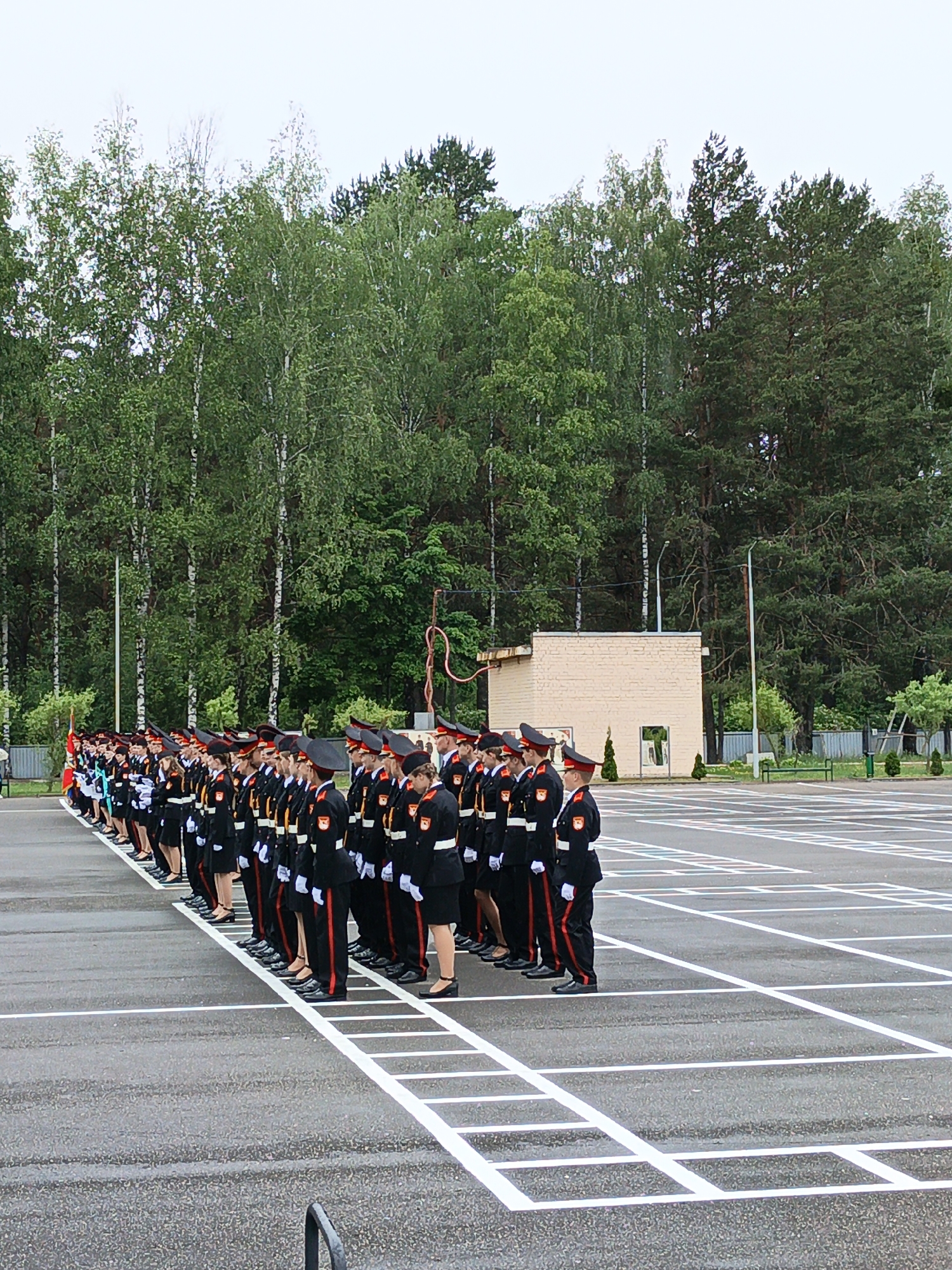 A group of cadets in uniform are standing in formation on an asphalt surface marked with white lines. The cadets are wearing black jackets with red accents and matching pants, along with red-trimmed caps. They are lined up in rows, facing towards the right of the frame. In the background, there is a low, light-colored brick building and a dense line of trees, primarily birches and pines. The sky is overcast. This scene appears to be outdoors, possibly in a parade ground or training area within the city of Porechye, Belarus.