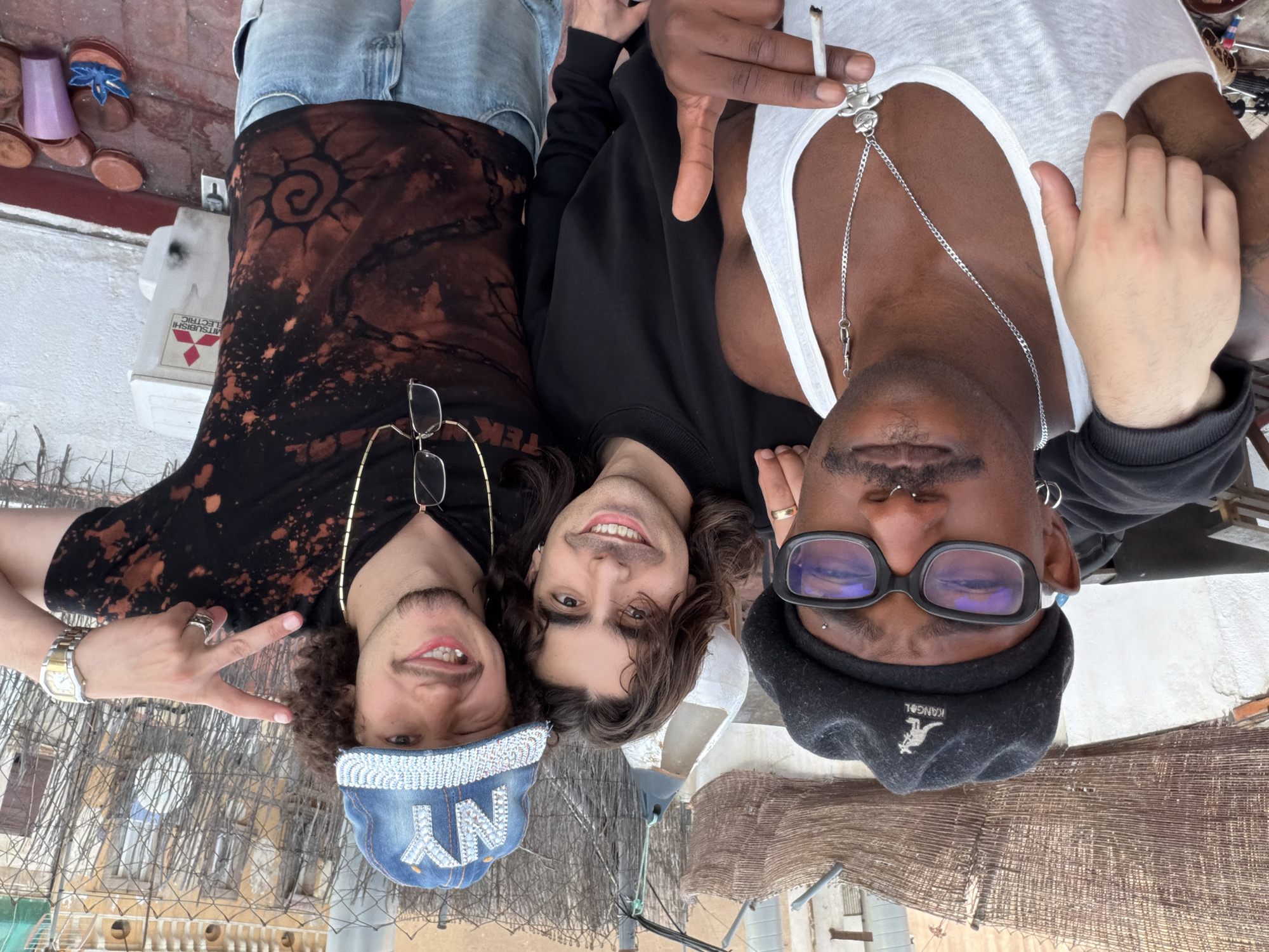Three men are posing for a photo on a rooftop in Barcelona. The man on the left is wearing a tie-dye shirt, blue jeans, and a baseball cap that says "NY" and is bedazzled. He is wearing glasses and a gold chain and is holding up a peace sign. The man in the middle is wearing a black sweatshirt and has shoulder-length brown hair. The man on the right is wearing a white tank top, black Kangol hat, and large black glasses. He has a cigarette in his hand and a silver chain around his neck. The background shows rooftops and a fence, and some buildings. The sky is overcast. A Mitsubishi Electric air conditioning unit sits at the edge of the roof.