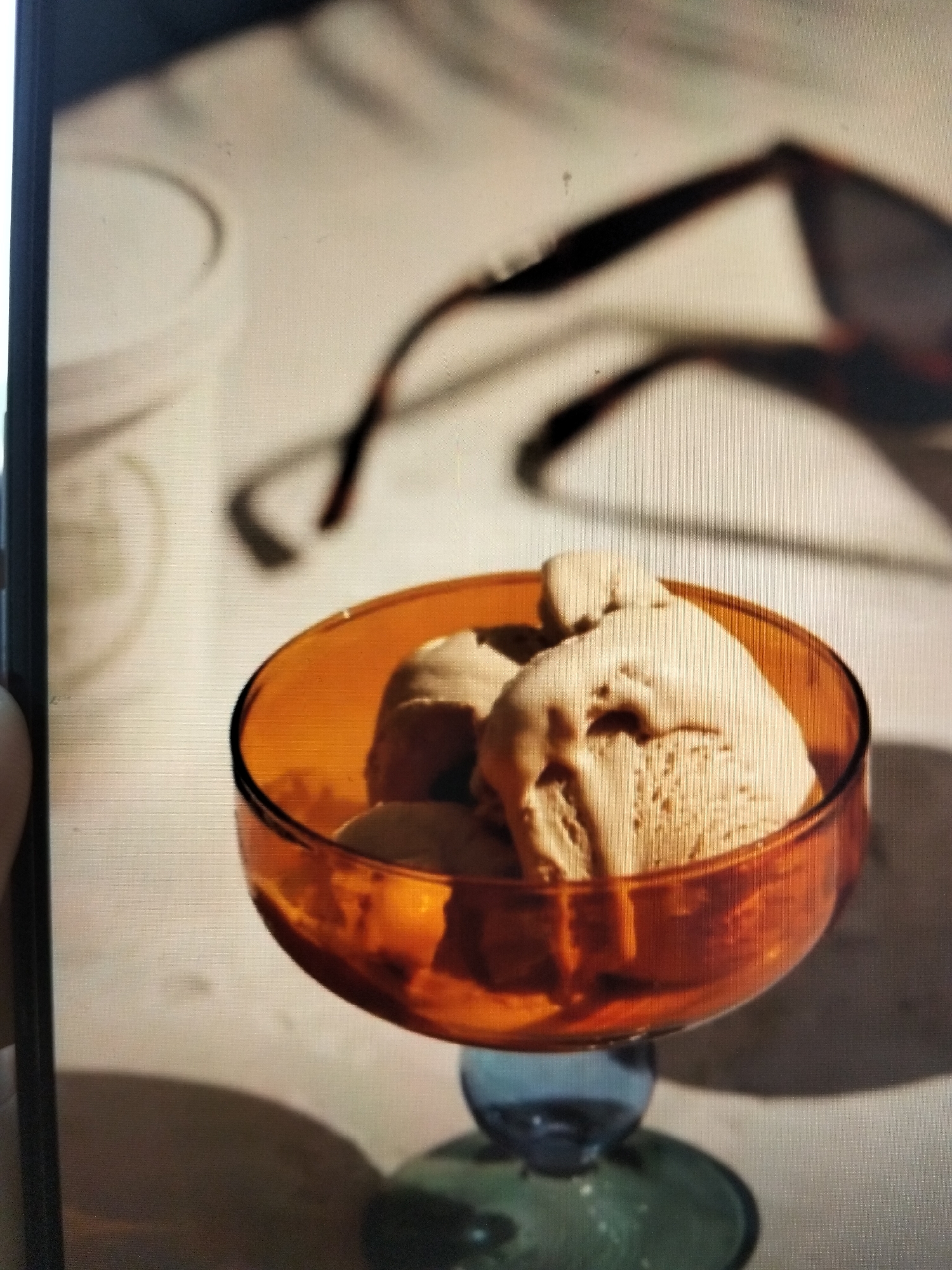 An amber glass dessert bowl with a blue and green stem holds several scoops of light brown ice cream. The bowl sits on a light-colored surface, possibly a table, which is partially in shadow. In the background, out of focus, are what appear to be sunglasses casting a shadow, and a white cylindrical container, possibly an ice cream tub. The scene is lit by a strong light source, creating distinct shadows. There are no people or animals present in the image. The location is not specified.