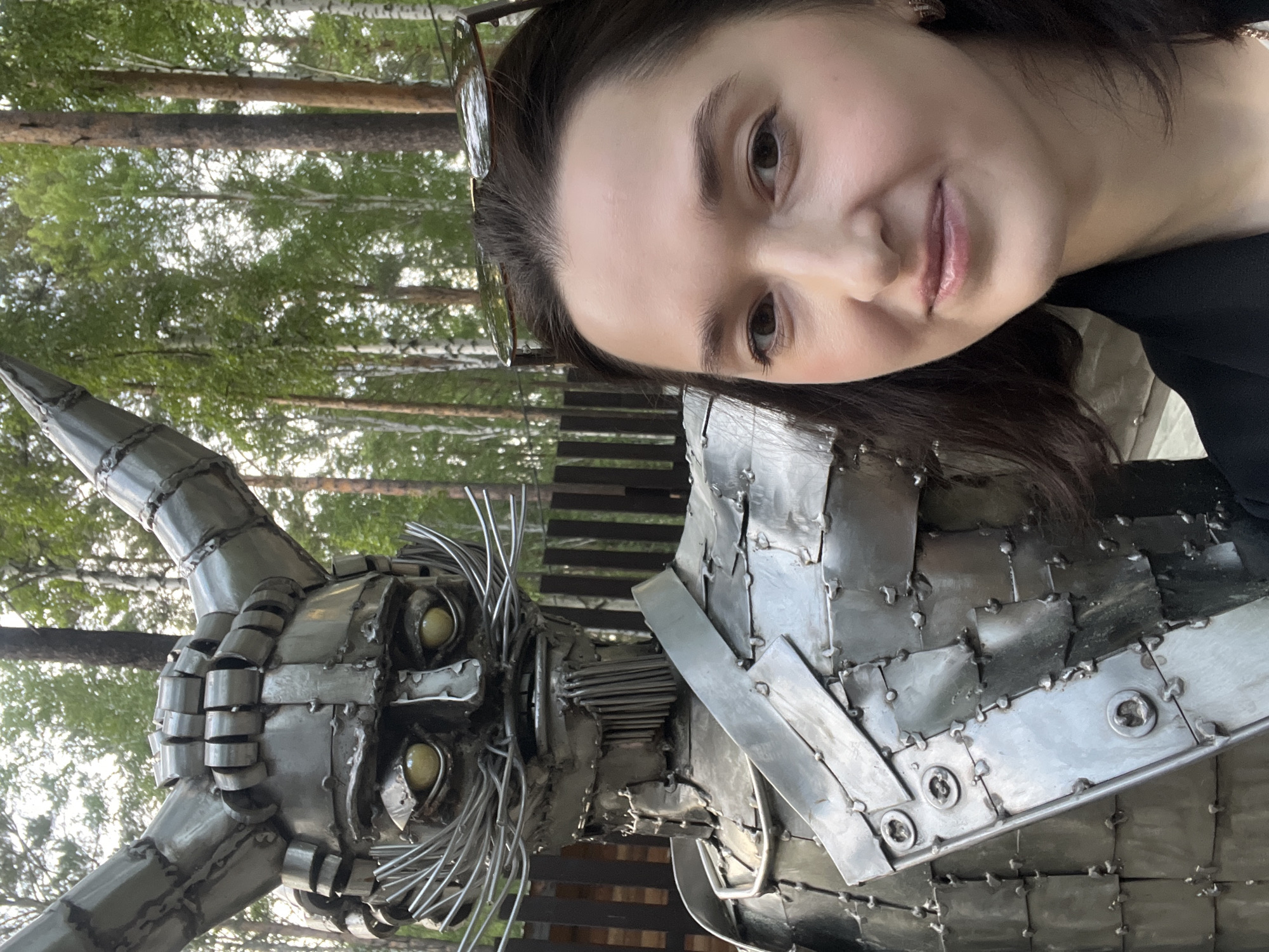 The image shows a woman taking a selfie in front of a large metal sculpture of a horned figure with a mustache, possibly a depiction of a character from folklore. The sculpture is made of many metal plates welded together, creating a segmented, armored appearance. The woman is in the foreground, smiling slightly and wearing a black jacket. Behind them, a wooden fence is visible, and beyond that, a forest with birch trees. The setting appears to be outdoors, in a natural environment. The location is specified as the Tal'tsy Museum of Wooden Architecture, Russia.