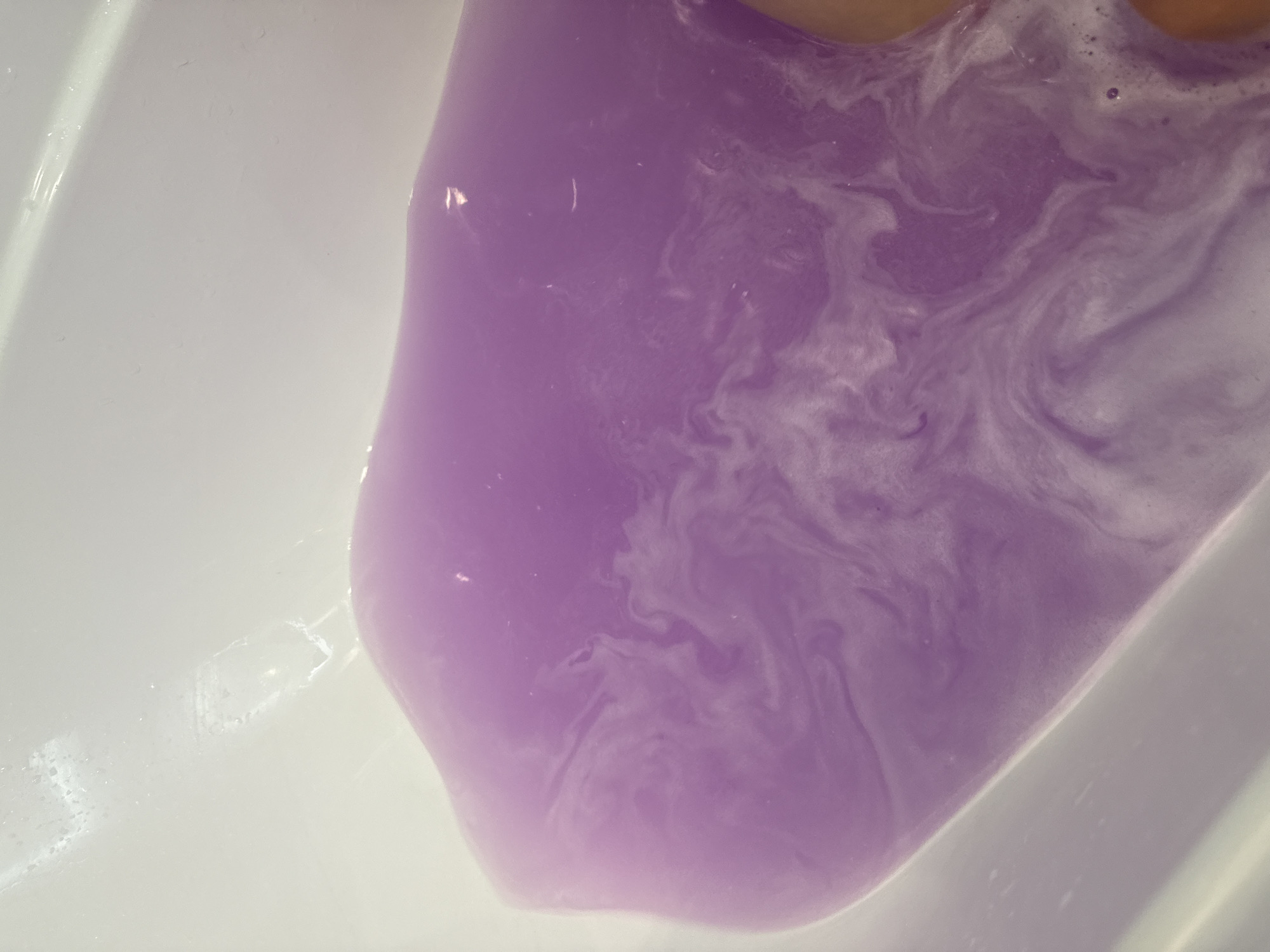 A close-up, overhead shot captures a bathtub filled with pale purple water. The water's surface exhibits swirling patterns of a lighter hue, suggesting the presence of bath products or movement. The white porcelain of the bathtub is visible along the edges, with some water droplets and slight imperfections. A portion of a person's leg, peeking in from the right side of the frame, indicates the bathtub is in use. The scene is indoors, likely a bathroom, and there is no specific indication of its location within Lyubertsy, Russia.