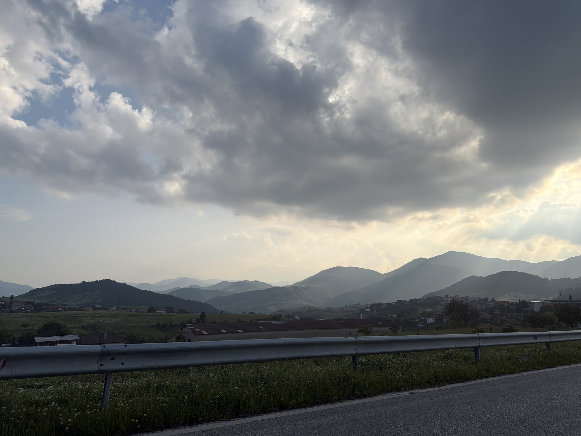 The image captures a panoramic view of a mountainous and rural landscape in Komuniga, Bulgaria, during what appears to be late afternoon or early evening. The sky is predominantly cloudy, with a mix of light and dark grey stratocumulus clouds dominating the upper portion. Patches of clear blue sky are visible on the far left. Towards the right, dense grey clouds are illuminated from behind by bright sunlight, casting visible light rays onto the landscape and creating a warm glow near the horizon.
In the immediate foreground, a dark grey asphalt road runs horizontally. Parallel to the road, a silver metal guardrail with white reflective markers separates the road from a grassy verge. The verge is covered in green grass and scattered small white wildflowers.
Beyond the guardrail, the midground and background consist of multiple layers of mountains and hills receding into the distance, characterized by atmospheric haze. The closest hills are dark green with visible vegetation. Several settlements are interspersed within this landscape. On the left, a small cluster of buildings with white walls and red roofs is visible. Towards the center-right, a large, elongated building with a reddish-brown roof is situated in a green field. Further to the right, a denser village or small town with numerous houses, featuring various roof colors, is nestled among the foothills and valleys. The settlement is partially illuminated by the sunlight breaking through the clouds. No people, animals, or vehicles are present in the visible scene.