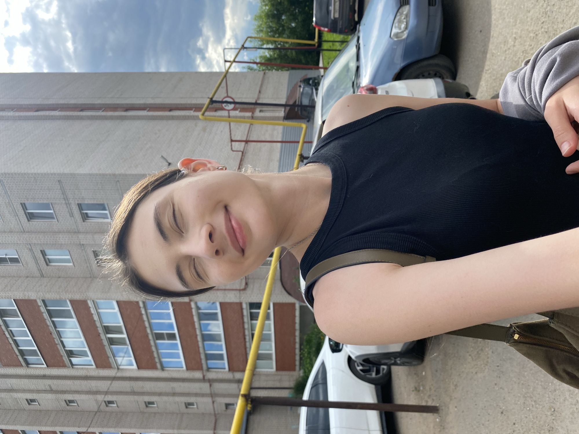 A young woman with closed eyes is taking a selfie outdoors in Votkinsk, Russia. She is wearing a black sleeveless top and has a bag slung over her shoulder. Behind her, a multi-story brick apartment building stands tall, with several cars parked in the foreground. The sky is partly cloudy.