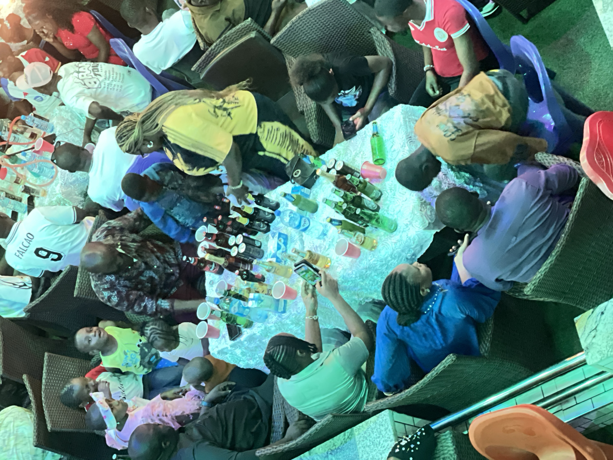 A gathering of approximately 15 people, including several children, are congregated around a table laden with bottles of alcoholic beverages and red plastic cups. The people are seated in wicker chairs and are dressed in a variety of casual clothing. One person is holding up a cell phone to take a photograph of the table. The location is Obosi, Nigeria.