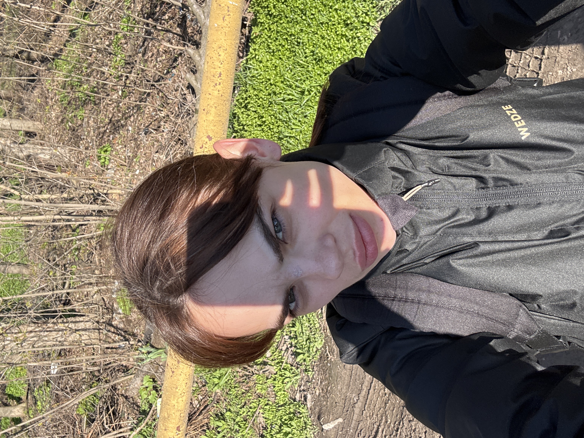 The image is a selfie of a young woman with brown hair and fair skin. She is wearing a black jacket with the word "WEDZE" visible on the right side of the jacket. The background consists of vegetation, including green grass, trees with bare branches, and a yellow metal pole. The ground appears to be a mix of dirt and concrete. Light and shadow play across her face, with distinct lines of sunlight. The location cannot be confirmed, but the prompt hints the city of Rostov-on-Don, Russia.