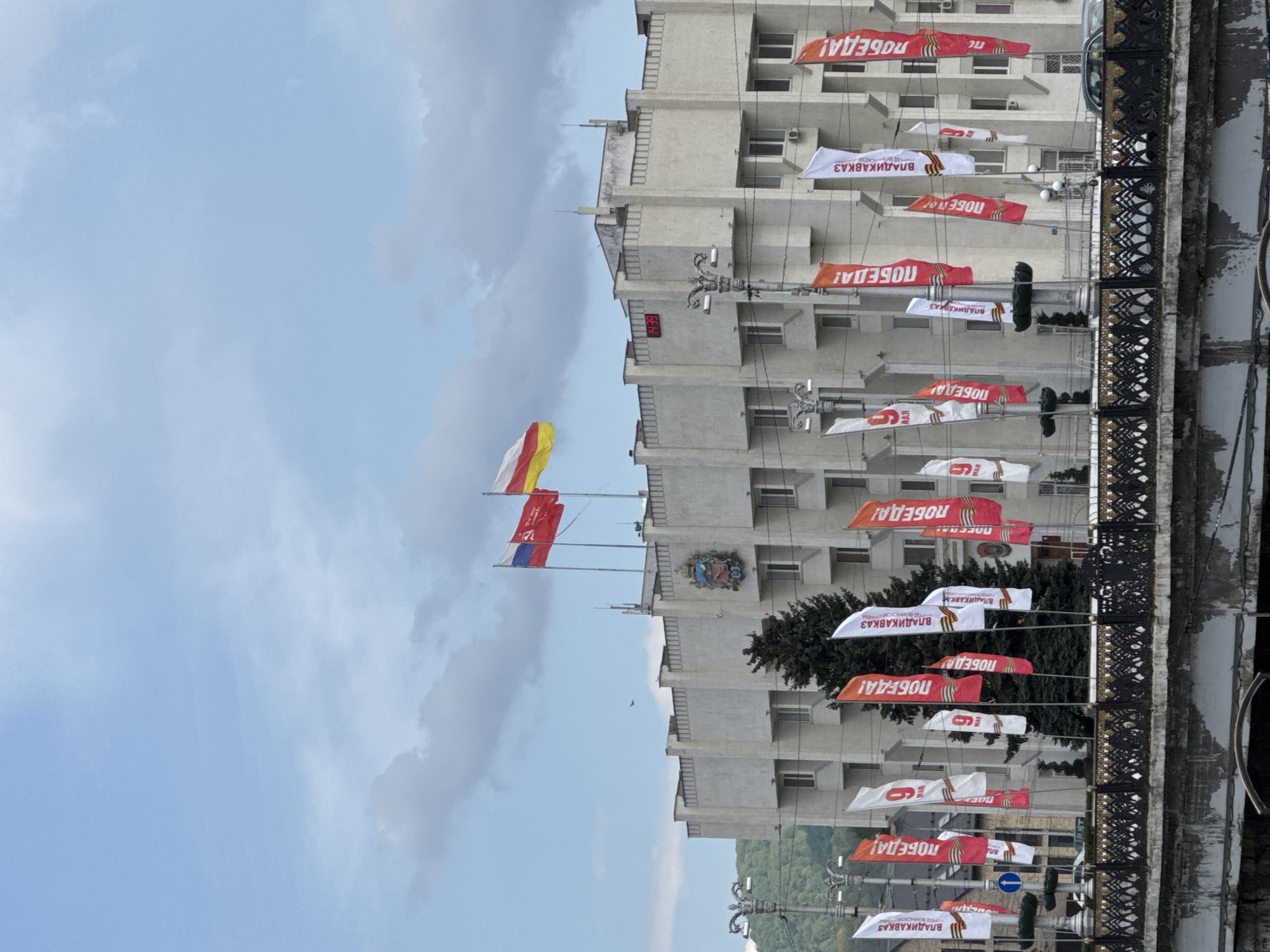 The image shows a building in Vladikavkaz, Russia, likely a government building, adorned with Russian and regional flags, and banners celebrating Victory Day. The building is a multi-story concrete structure with repetitive window patterns. Several flagpoles are present, displaying the Russian flag, the flag of North Ossetia–Alania, and another red flag. Banners strung between lamp posts along the street proclaim "Victory!" in Russian ("ПОБЕДА!"). Some banners also display "Vladikavkaz - Hero City" (ВЛАДИКАВКАЗ ГОРОД ВОИНСКОЙ СЛАВЫ). The sky is partly cloudy. A bridge with intricate iron railings is visible in the foreground, possibly reflecting in a body of water. There are hints of greenery, possibly trees, interspersed between the building and the bridge. The building features a coat of arms.