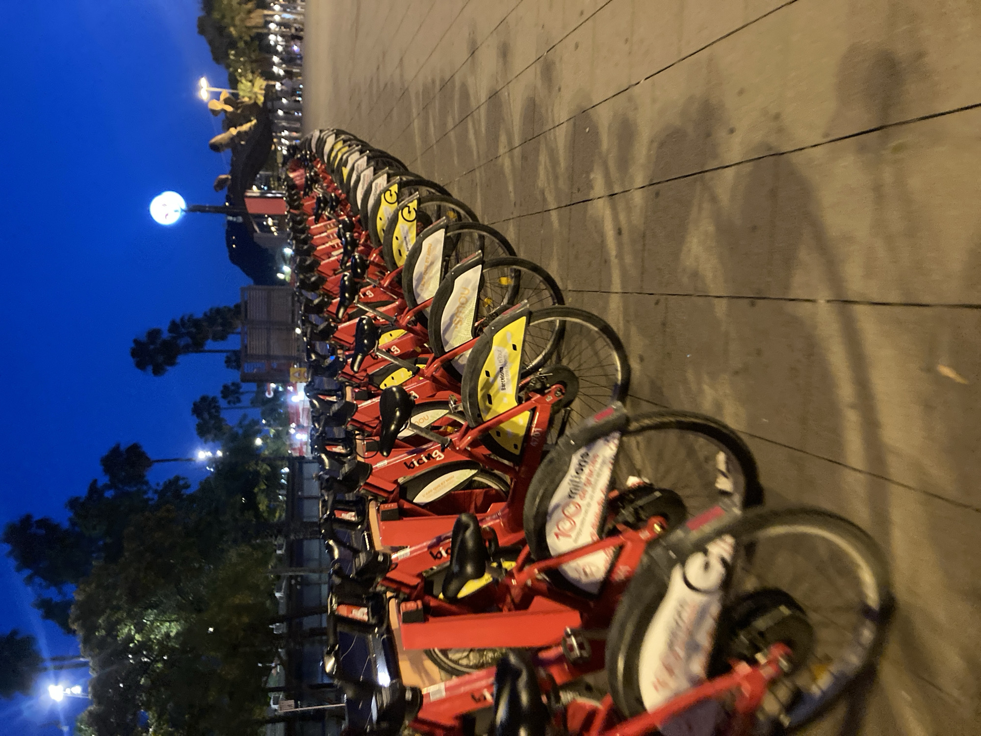 The image depicts a row of red bicycles parked along a paved walkway in Barcelona, Spain, at dusk. The sky is a deep blue, transitioning from twilight to night, with a bright circular light visible in the distance, possibly a street lamp or a decorative element. Palm trees and other foliage are silhouetted against the sky, suggesting a Mediterranean climate. The bicycles are uniform in design, featuring red frames, black seats, and yellow accents on the wheels. Some of the bicycles have white fenders with text, including "100 millions" and "Bicing," which is the name of Barcelona's public bicycle-sharing system. The walkway is made of large, rectangular paving stones, and some bicycles cast shadows on it. The scene appears to be in an open public area, possibly a promenade or a plaza. There are no people visible in the immediate foreground or among the bicycles. In the background, faint lights suggest the presence of buildings and streetlights, indicating an urban environment. The overall impression is one of a quiet, orderly urban scene at the end of the day.