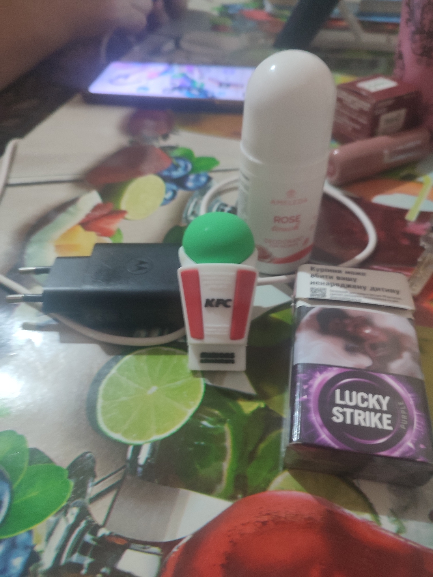 The image depicts a close-up, overhead view of a table displaying various items. In the foreground, a pack of Lucky Strike cigarettes with purple packaging is visible. To the left of the cigarettes, a black Motorola phone charger is present. Further to the left, a small white and red KFC container with a green top sits on the table. In the background, a white roll-on deodorant with "Amela" and "Rose" written on it is visible. A blurred smartphone screen with colorful graphics is also present. The table surface appears to be a placemat with a printed fruit design, including images of limes, strawberries, and blueberries. The lighting suggests an indoor setting, possibly a home or office environment. There is no discernible information or context within the image to indicate its specific location in Chernivtsi, Ukraine.