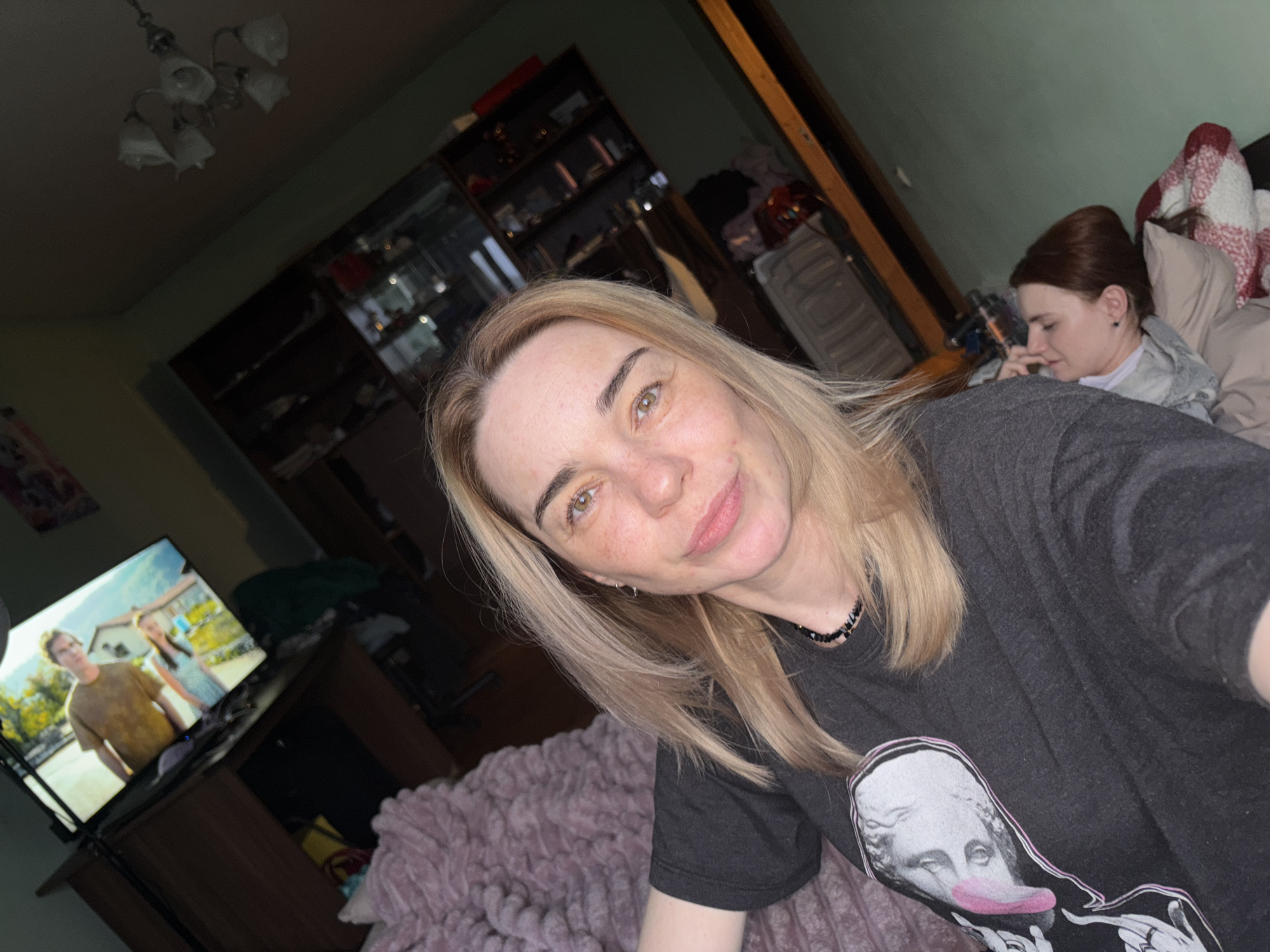 A selfie-style photo captures two women relaxing indoors, likely in a living room or bedroom, during the evening or night.
In the foreground, a woman with blonde hair and light-colored eyes smiles gently at the camera. She has light freckles and is wearing a dark gray t-shirt featuring a stylized graphic of a classical bust with a pink tongue sticking out and hands making a "rock on" gesture. She also wears a black beaded necklace and a small hoop earring. She is positioned over a fluffy pink or purple blanket.
In the upper right background, another woman with reddish-brown hair is partially visible, lying down on a bed or couch, seemingly resting or engaged with a device out of frame. She is covered by a red and white plaid blanket.
To the left, a flat-screen television displays a scene with two people (a man and a woman) outdoors, suggesting a show or movie is playing. The TV sits on a dark wooden stand. Overhead, a white, multi-armed chandelier with flower-shaped shades is visible. The room has dim lighting, with visible furniture including dark shelving units or cabinets in the background, containing various items. A light-colored suitcase is also discernible near the second woman.