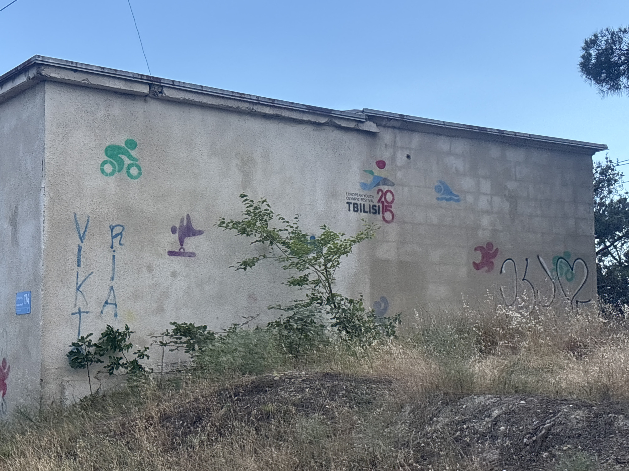 This media file depicts the exterior of a concrete building adorned with various graffiti and murals. The scene appears to be outdoors, with dry, tall grass in the foreground and the blue sky visible above. Trees are present to the right and behind the building.
The graffiti includes several stylized human figures engaged in athletic activities, such as cycling, swimming, and running. One prominent mural features the logo for the "European Youth Olympic Festival Tbilisi 2015," with the year prominently displayed. There is also text spray-painted vertically on the left side of the building, appearing to be in blue letters, though the full word or phrase is not entirely clear. On the right side, there is graffiti that includes a heart shape.
The lighting suggests it is daytime, likely sunny, given the clear blue sky and the way the sunlight illuminates the building and surrounding vegetation. The overall impression is one of an urban or semi-urban environment where public art has been applied to an older structure. The presence of the Olympic Festival logo provides a specific temporal and locational clue, indicating a connection to an event that took place in Tbilisi in 2015. The vegetation in the foreground appears dry and somewhat unkempt, suggesting a warm or dry climate.