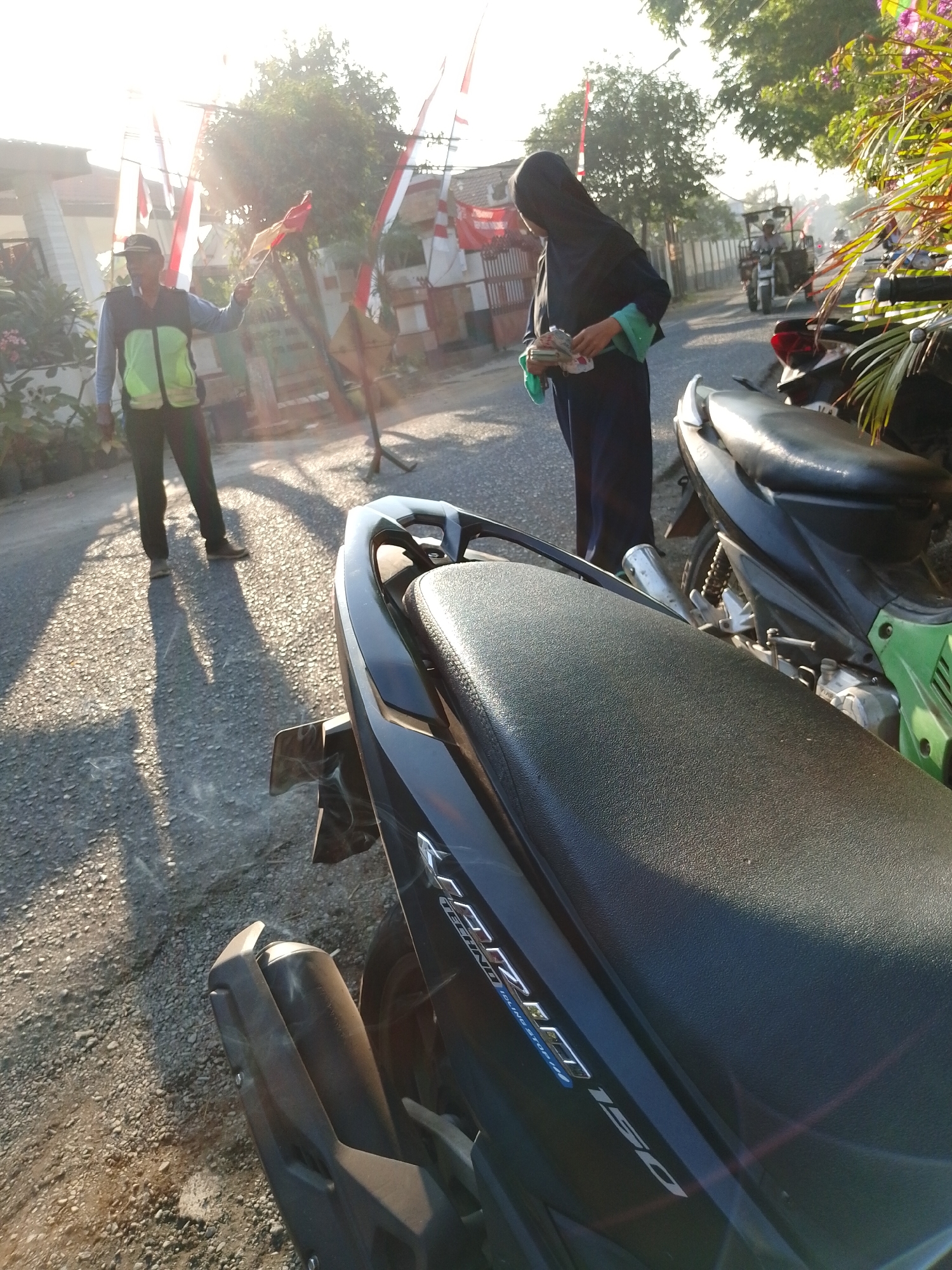 In Pasuruan, Indonesia, during daylight hours with bright sunlight casting long shadows, a man wearing a cap and a reflective vest is standing on a gravel road, holding a small Indonesian flag. To his right, a person in a dark hijab and long dark clothing is holding a bundle of money and a green cloth. In the foreground, the black seat of a motorcycle is visible, with a portion of its dark body and a silver muffler on the left. Further back, another motorcycle, mostly green, is parked. A tractor is moving down the road in the background, and several Indonesian flags on poles are lining the side of the road, suggesting a patriotic or celebratory occasion. The environment appears to be a semi-urban or rural area with houses and trees.