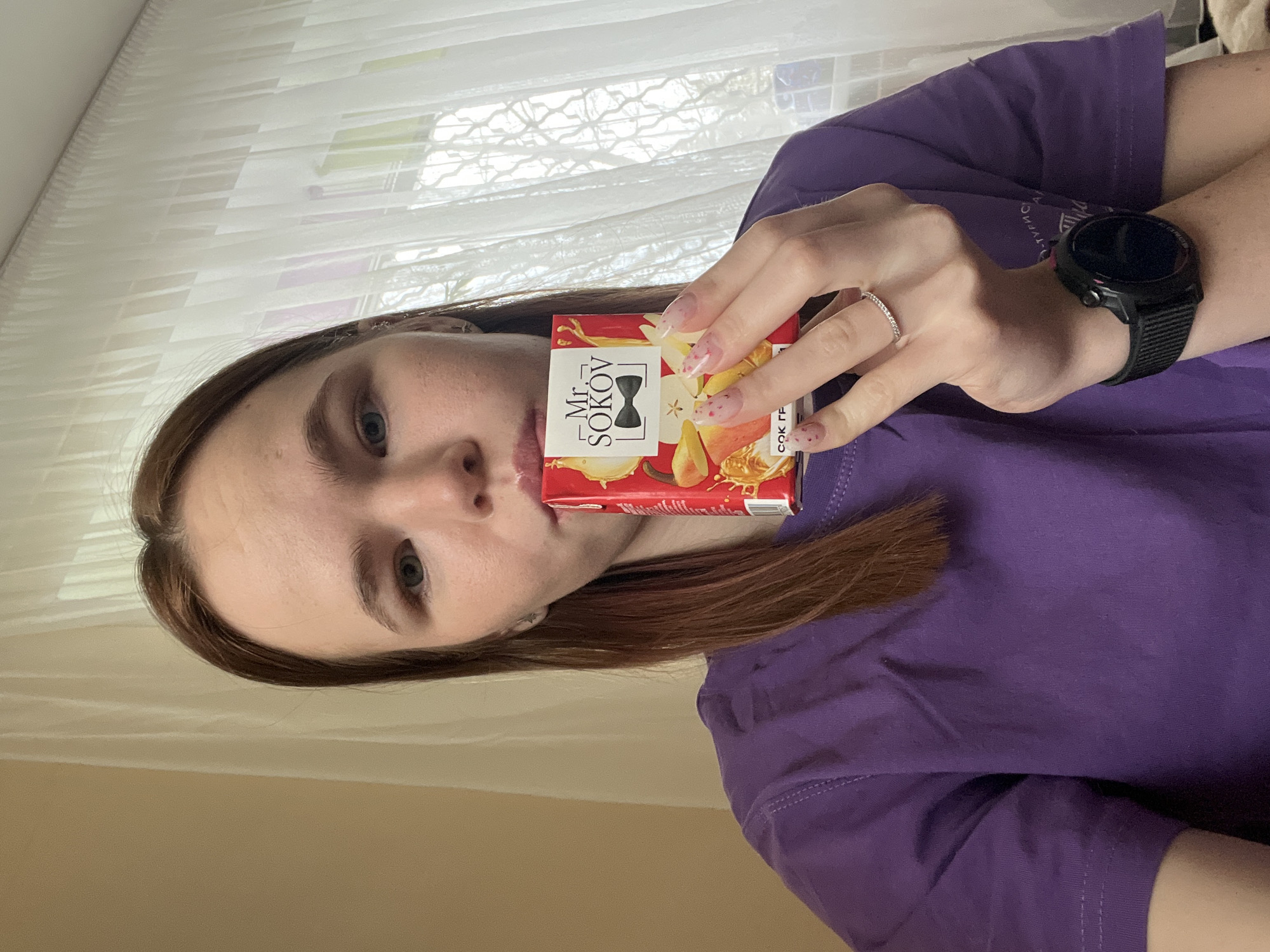 A young woman stands indoors, presenting a small, red and white juice box of "Mr. Sokov" brand. The juice box has a fruit illustration. The woman is wearing a purple t-shirt and a black smartwatch. She also has a diamond ring on her left ring finger. She has medium-length brown hair, fair skin, and appears to have light-colored eyes. The background features a window with sheer white curtains.
There is no information to definitively place the scene in the city of Berezniki, Russia based solely on the image.