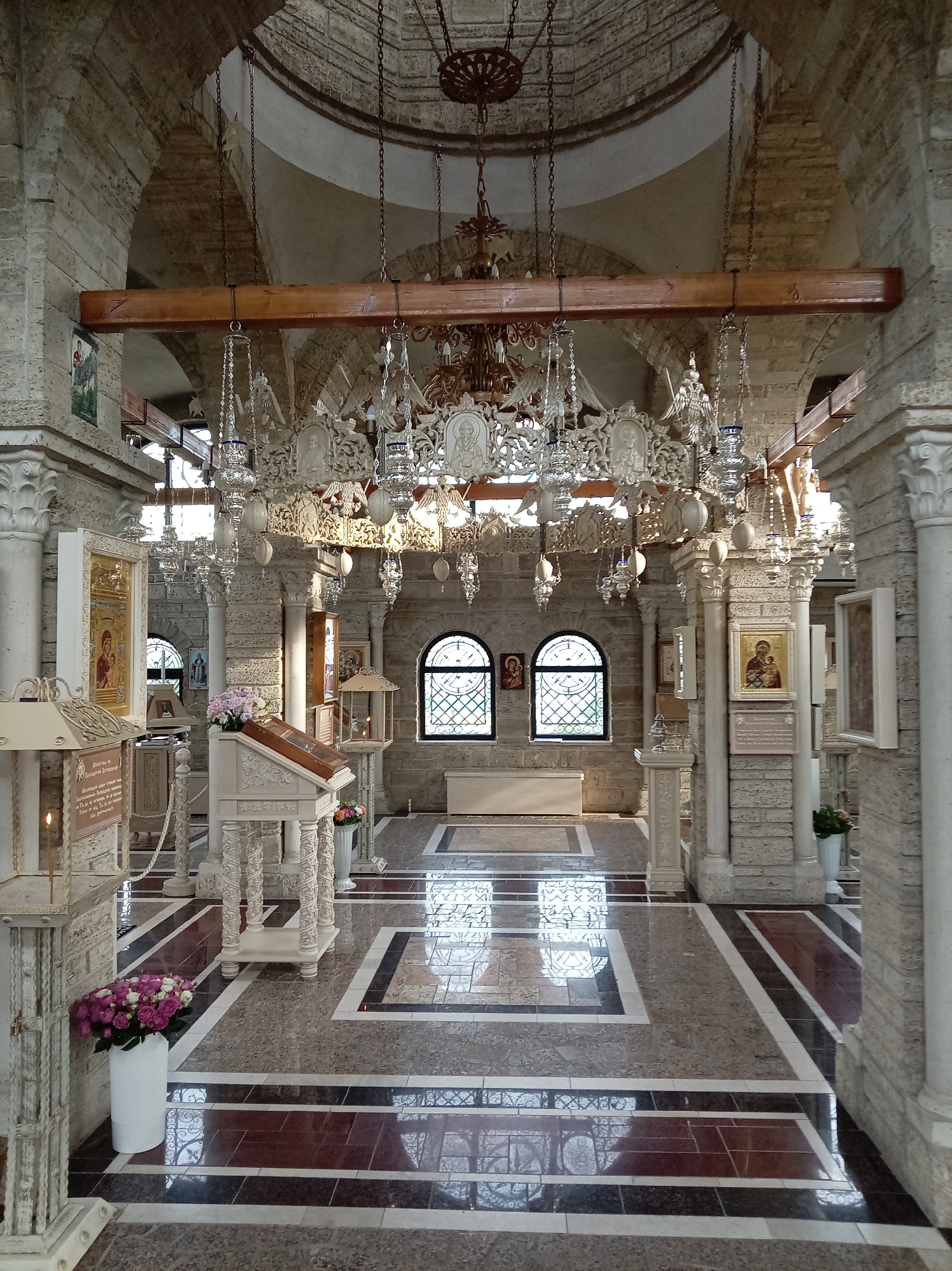 The image shows the interior of a stone church, likely the Ascension Cathedral in Yessentuki, Russia. The space features ornate chandeliers hanging from chains, stone pillars, arched ceilings, and a patterned tiled floor that reflects the light. Religious icons are displayed on stands and walls. In the foreground, a white vase holds a bouquet of pink and white roses. The scene appears to be peaceful and solemn, with no people visible. The architecture suggests a traditional Eastern Orthodox style.