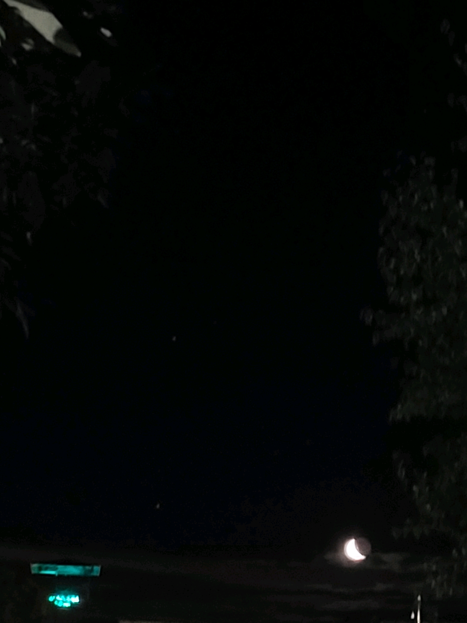 The image depicts a night scene in Ozolnieki, Latvia. In the foreground, silhouetted foliage frames the composition from the top left and right. A crescent moon is visible in the lower right quadrant, partially obscured by faint clouds. Below the moon, a faint horizontal structure with two glowing green lights is present, possibly indicating a street lamp or building signage. The sky is dark, with a few small, distant points of light that could be stars or other celestial bodies. The overall impression is a tranquil night sky with subtle man-made illumination.